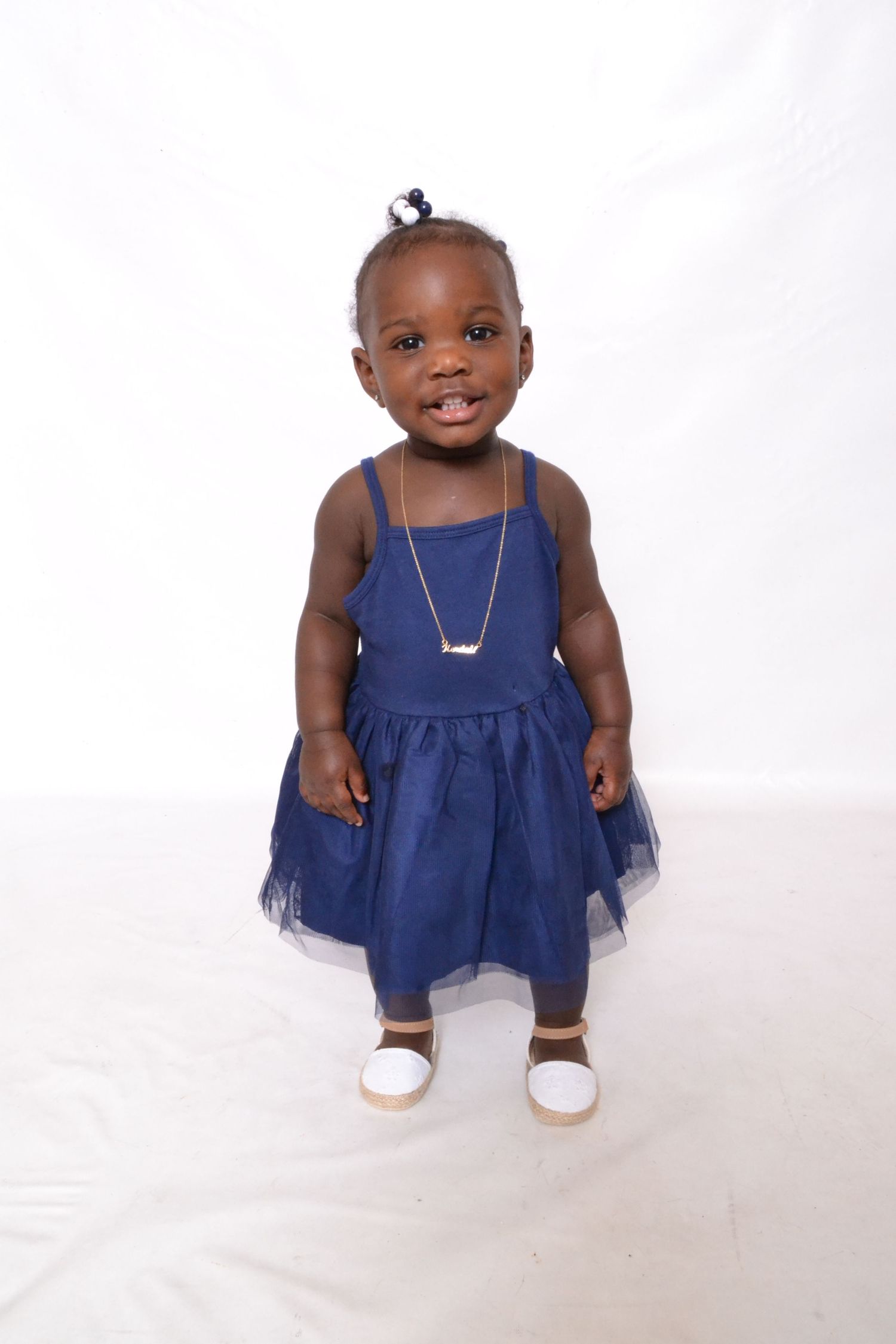 Smiling toddler girl standing in navy blue tutu dress and white shoes on white background.