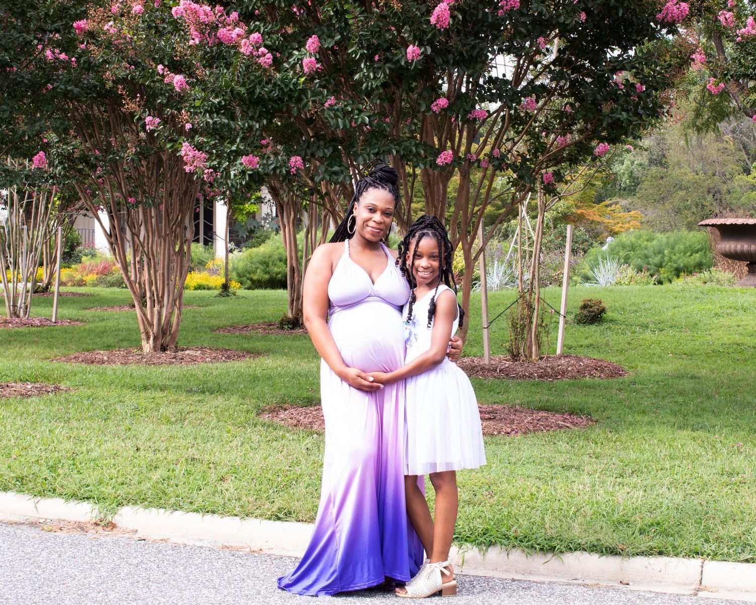 A pregnant woman in a purple ombre dress poses with a young girl in white near a flowering tree outdoors.