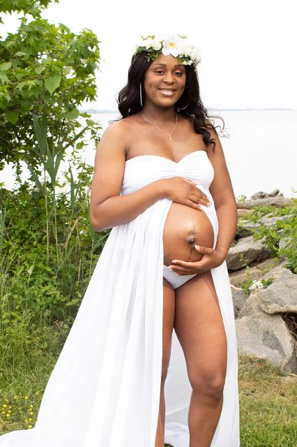 Pregnant woman in white dress with floral crown cradles baby bump outdoors near rocks and greenery.