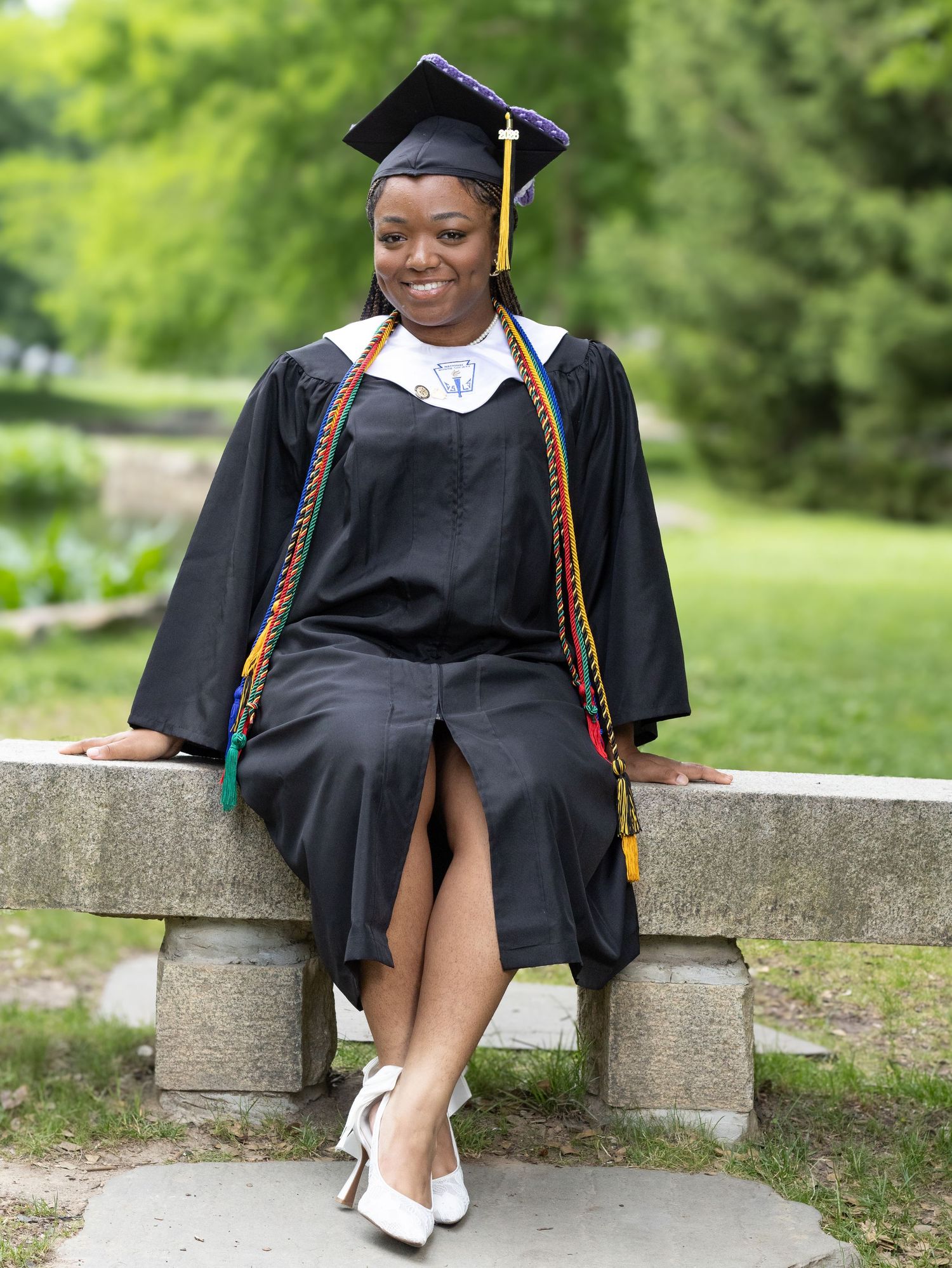 Graduate in black cap and gown with colorful honor cords sits on stone bench outdoors, smiling proudly.