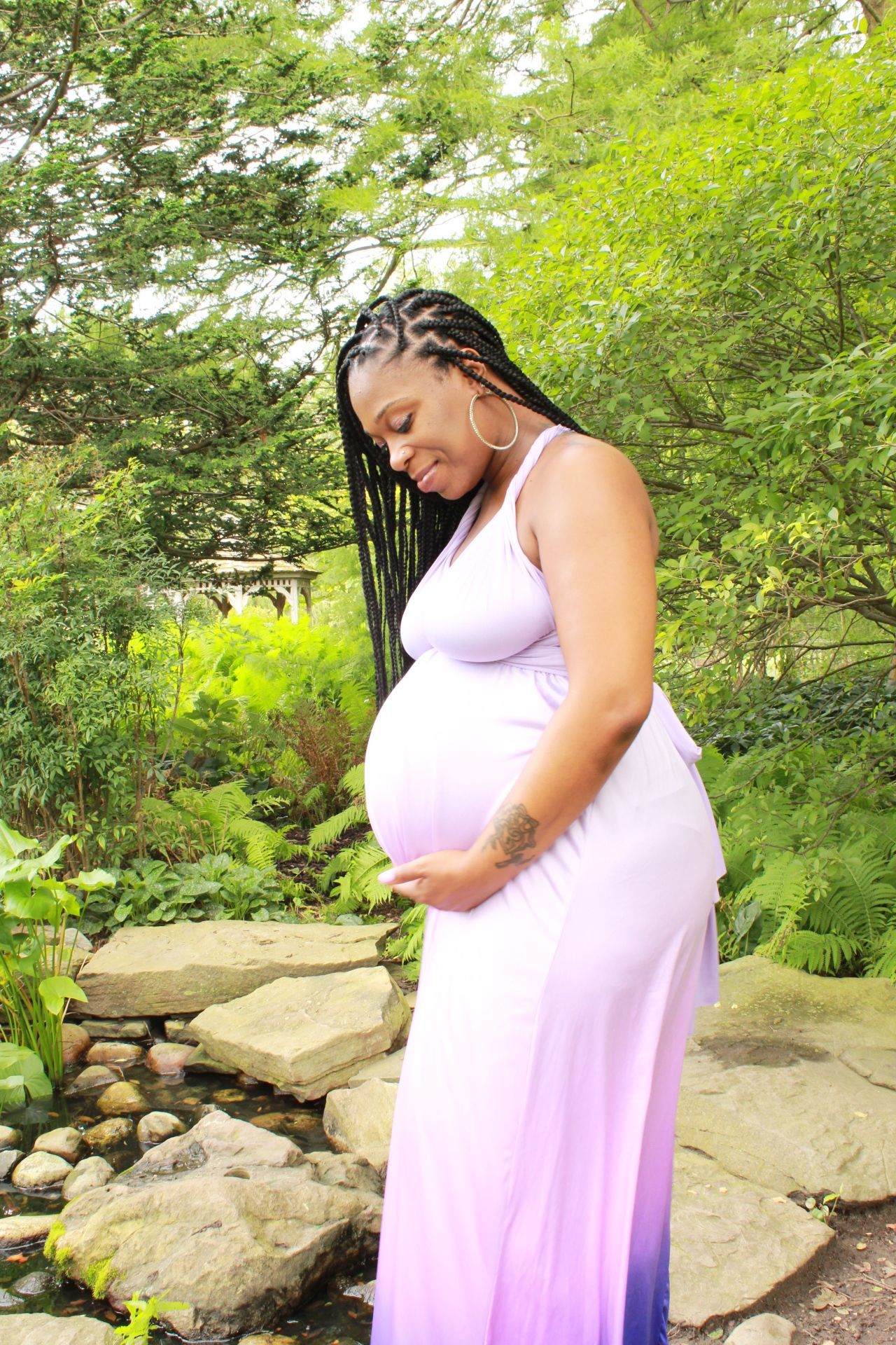 A pregnant woman in a lavender dress gently holds her baby bump while standing in a lush garden setting.