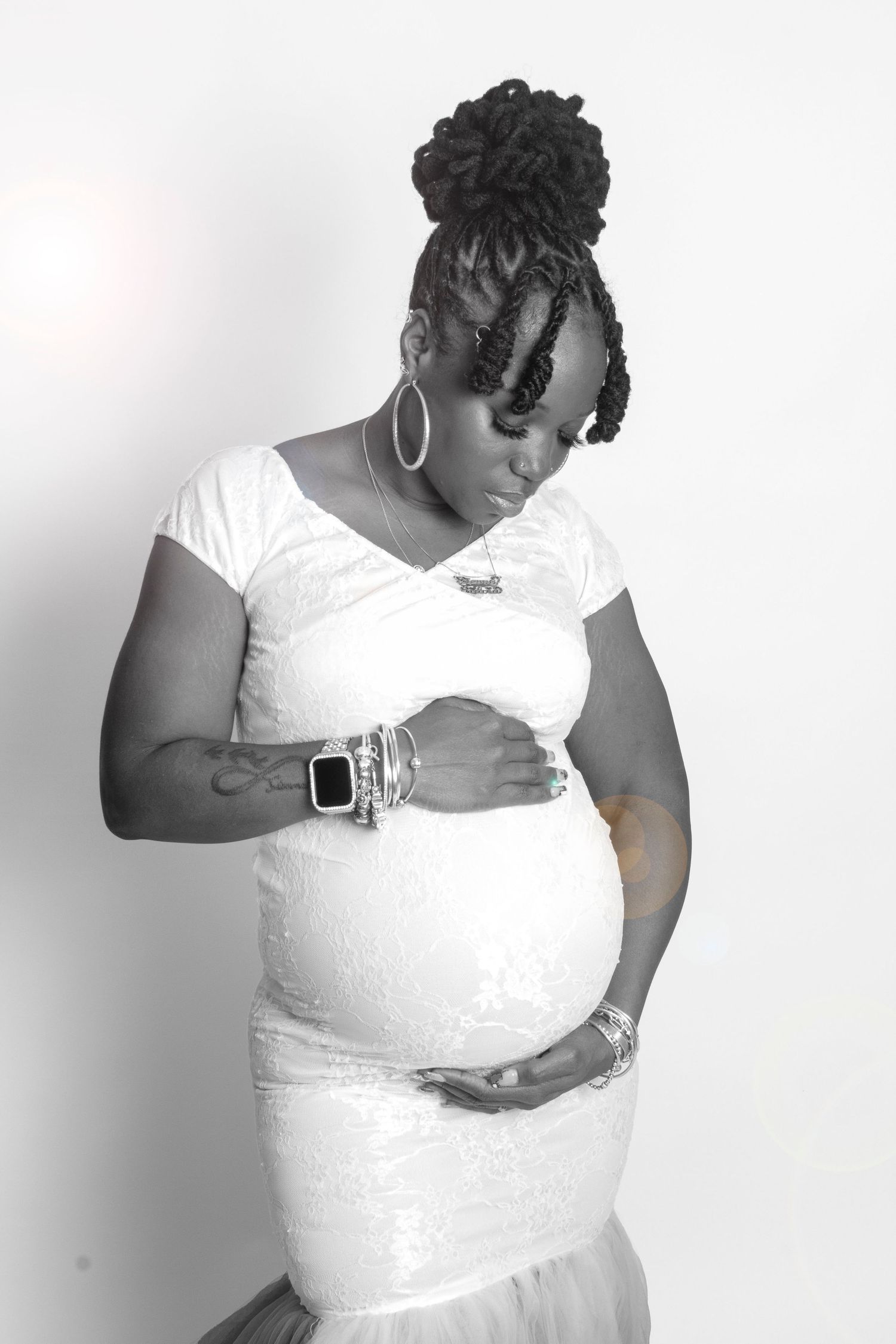 Black and white photo of pregnant woman in white lace dress with updo hairstyle cradling her baby bump.