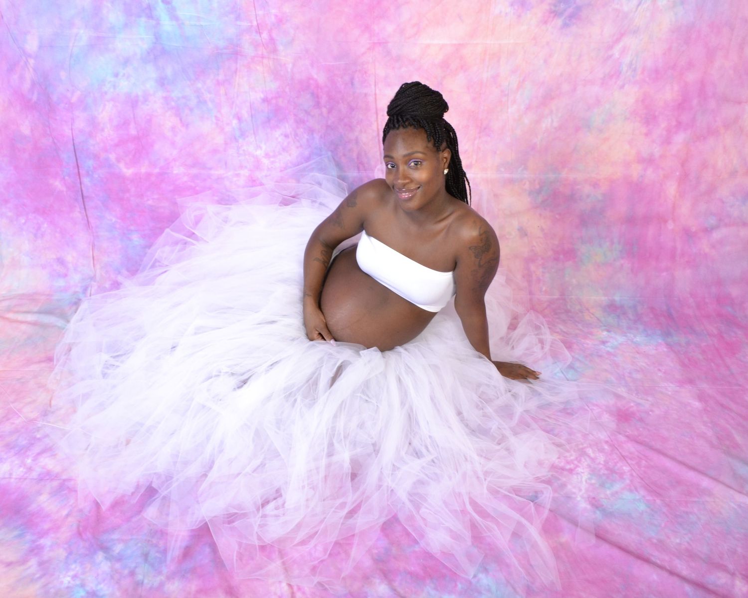 Pregnant woman in white tulle skirt sits gracefully against vibrant pink and blue tie-dye backdrop.