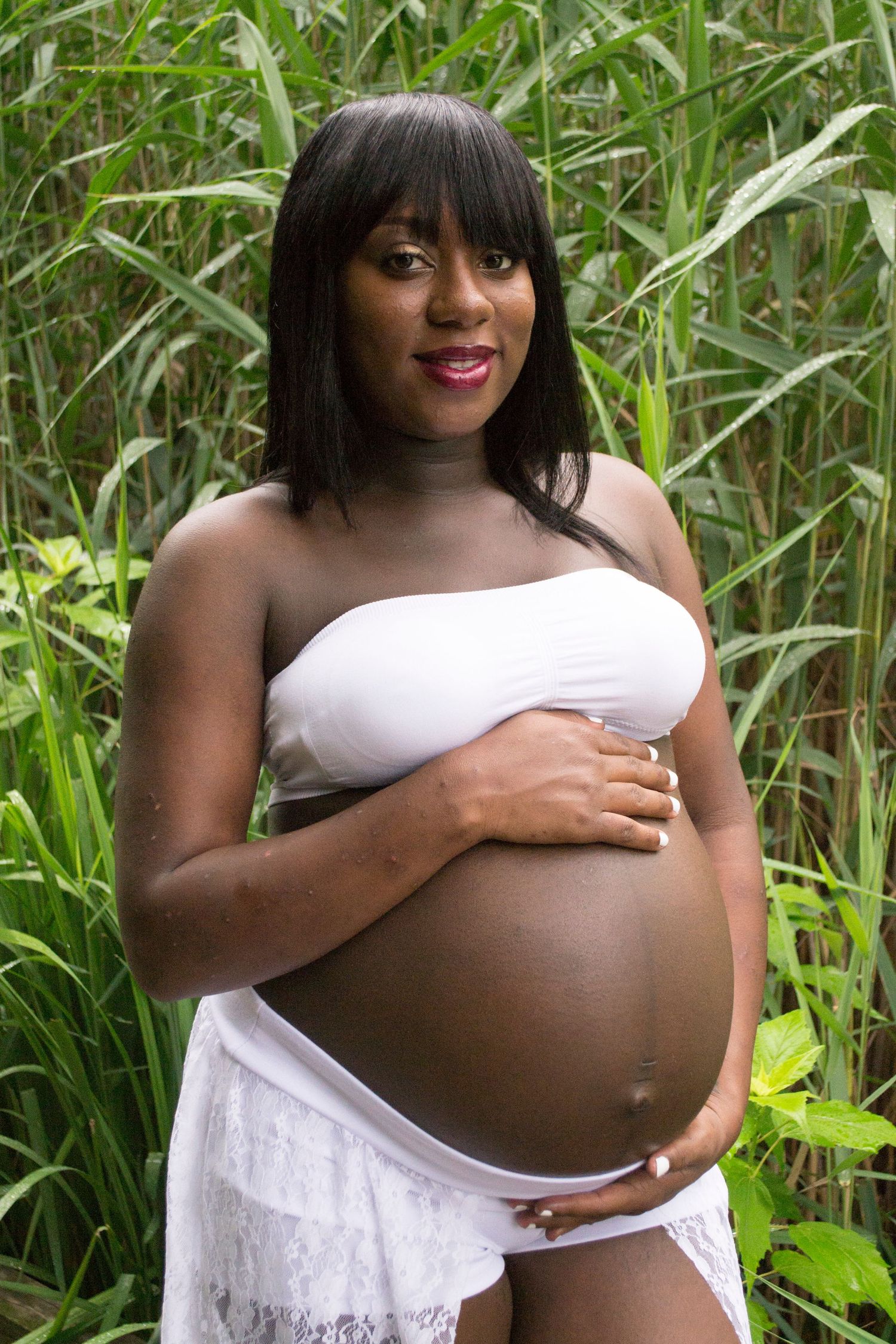 A pregnant woman in a white strapless outfit cradles her bare baby bump while standing among tall green reeds.