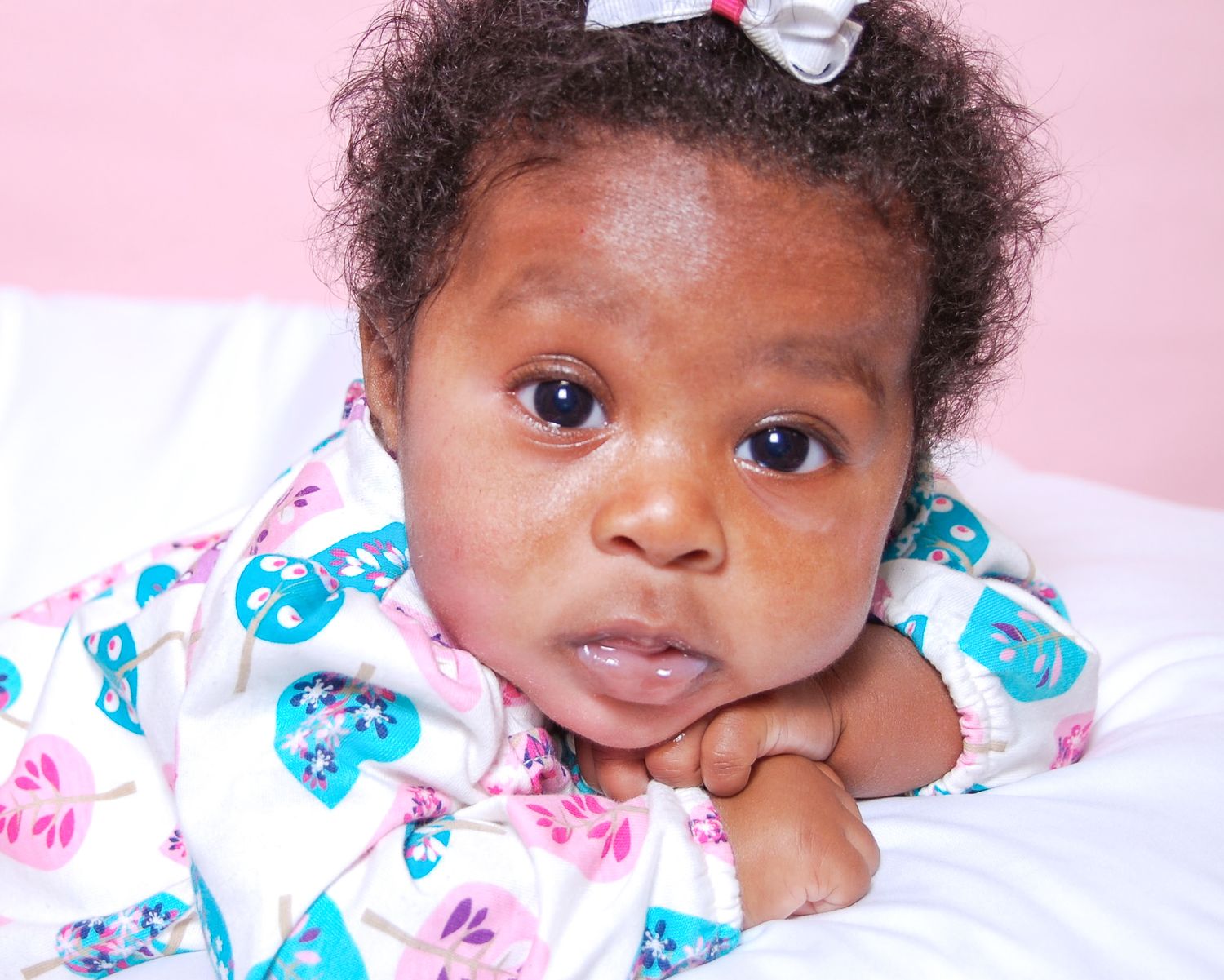 Adorable baby girl in floral outfit with white bow, lying on white surface against pink background.