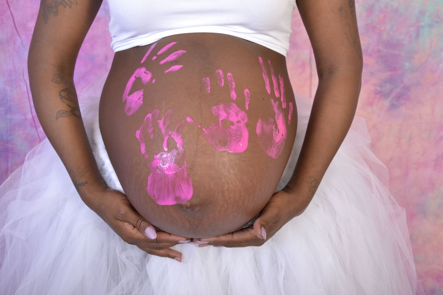 Close-up of pregnant belly adorned with pink handprint paintings on white tulle skirt background.