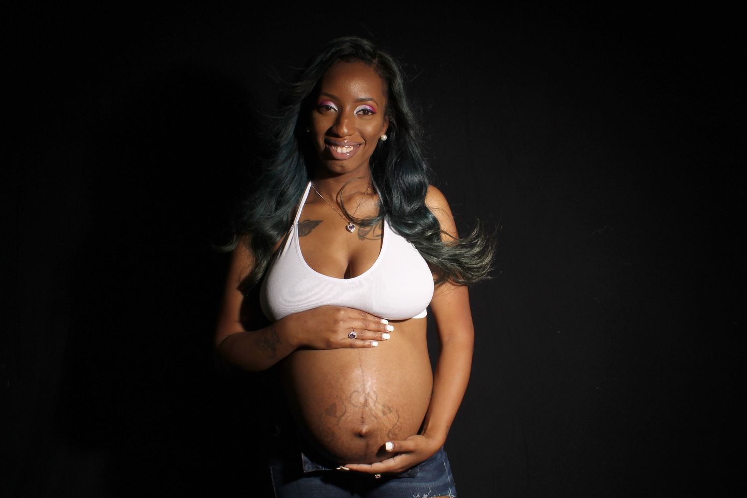 Smiling pregnant Black woman in white sports bra and jeans cradles her baby bump against dark backdrop.