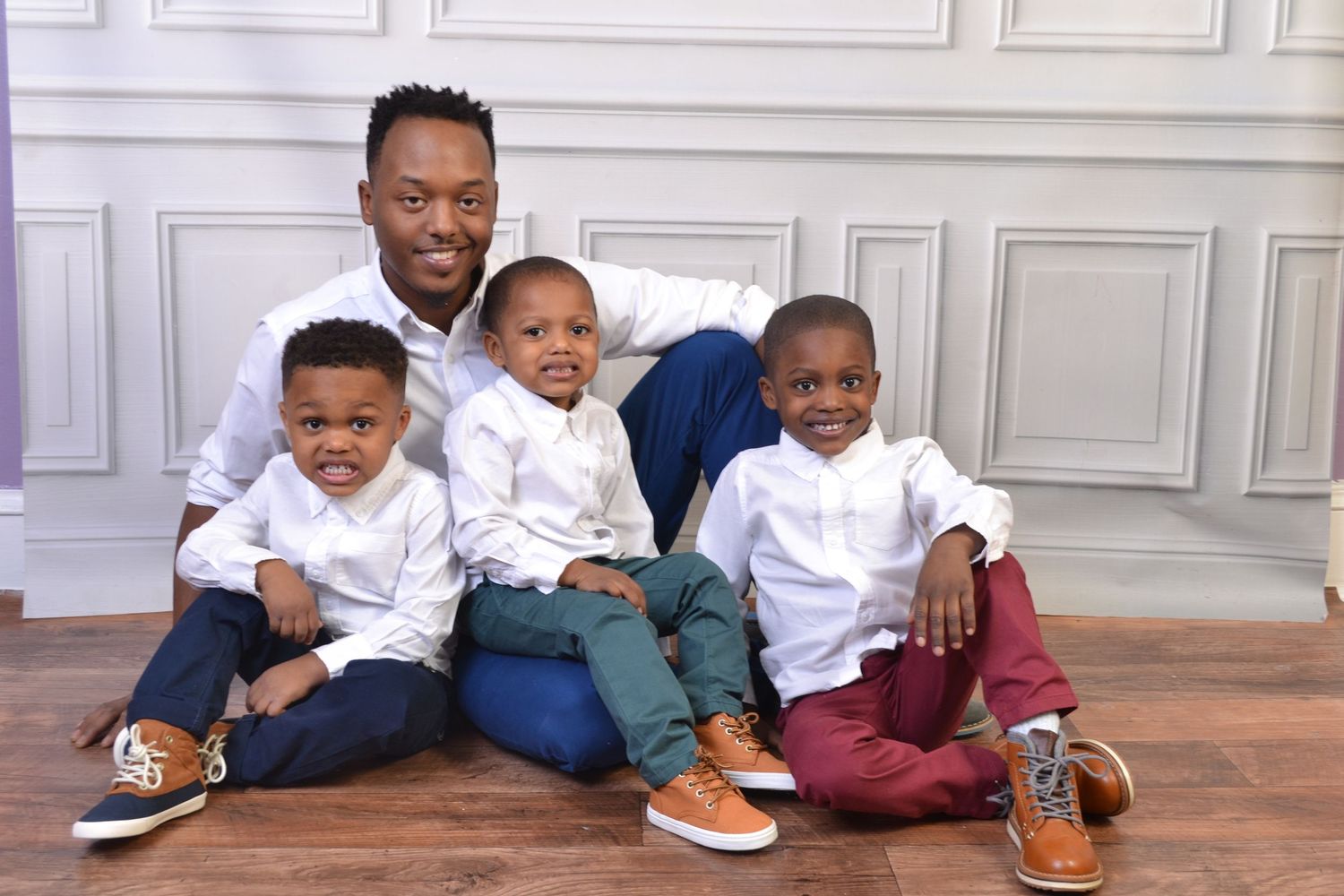 Father poses with his three young sons all wearing white shirts and brown loafers in a studio portrait.