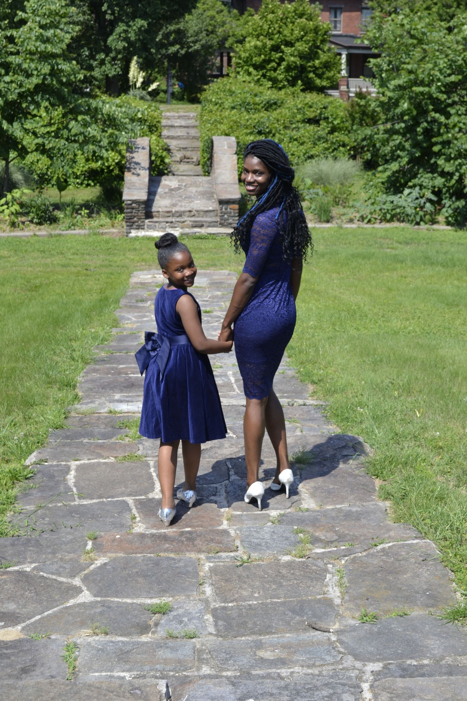 A woman and young girl in matching navy blue outfits hold hands while walking on a stone path in a park.