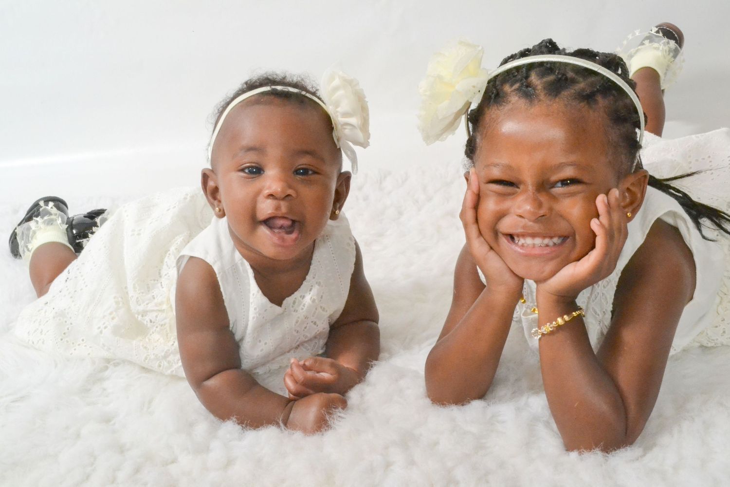 Two smiling young girls in white dresses with floral hair accessories lying on a fluffy white rug.