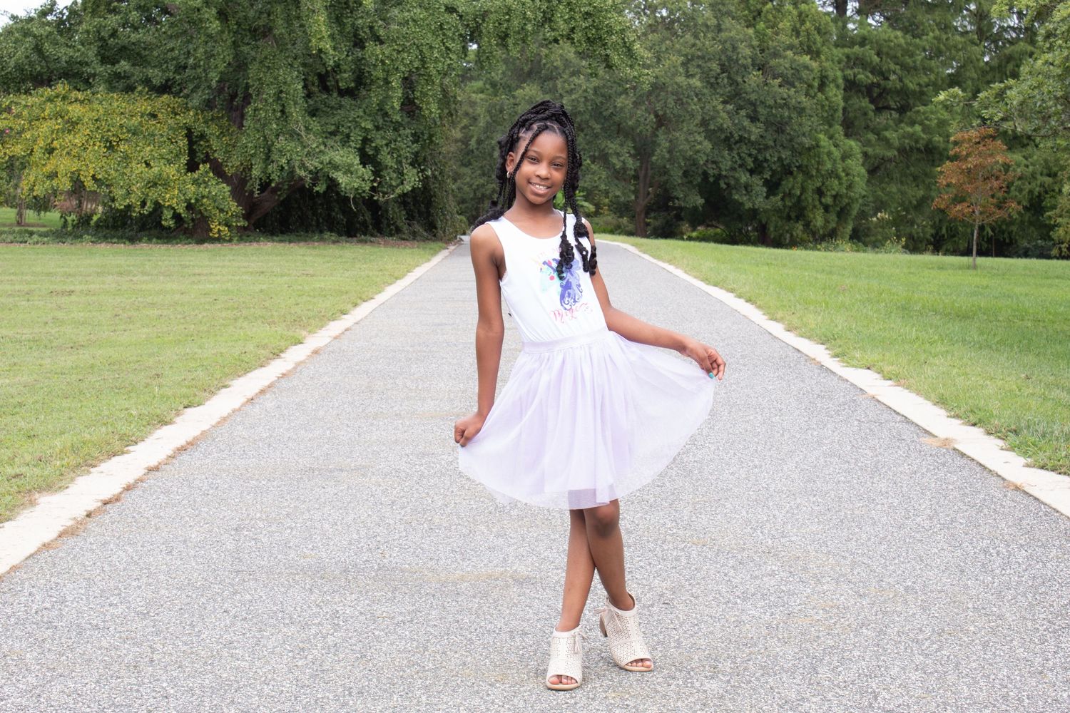 A young Black girl in a white tulle dress and heels poses gracefully on a park pathway surrounded by green trees.