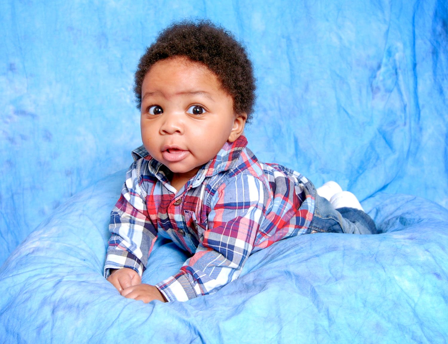 Adorable baby boy in plaid shirt poses on blue fabric backdrop for a professional portrait photo.