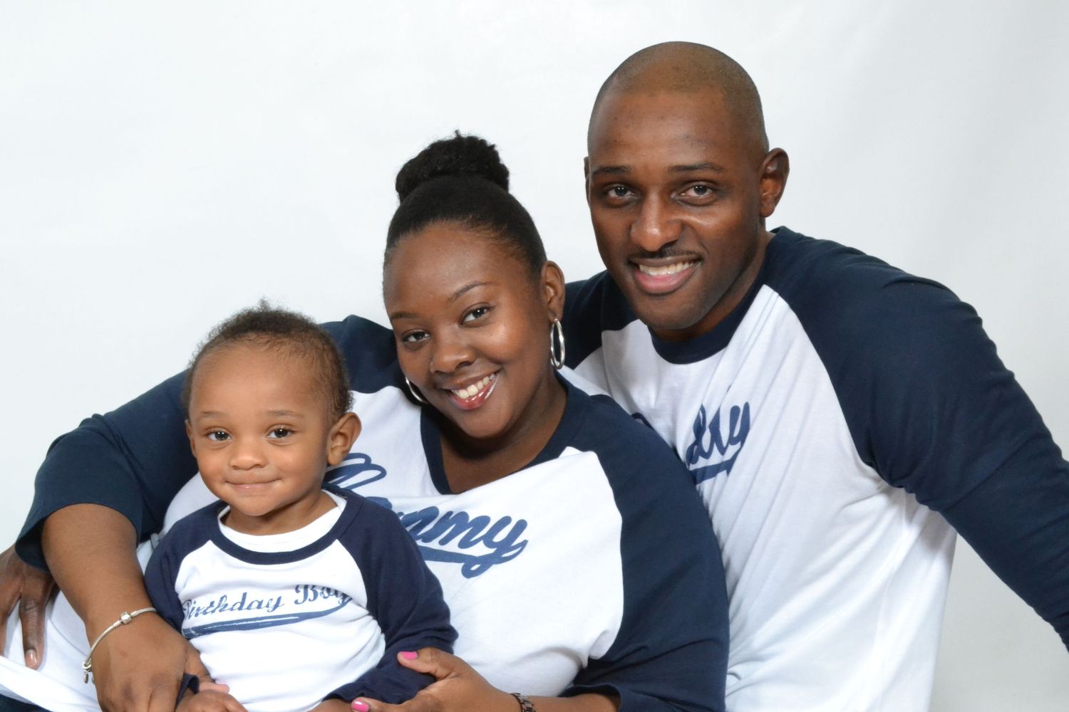 Happy young family of three wearing matching navy baseball shirts pose together in a bright studio portrait.