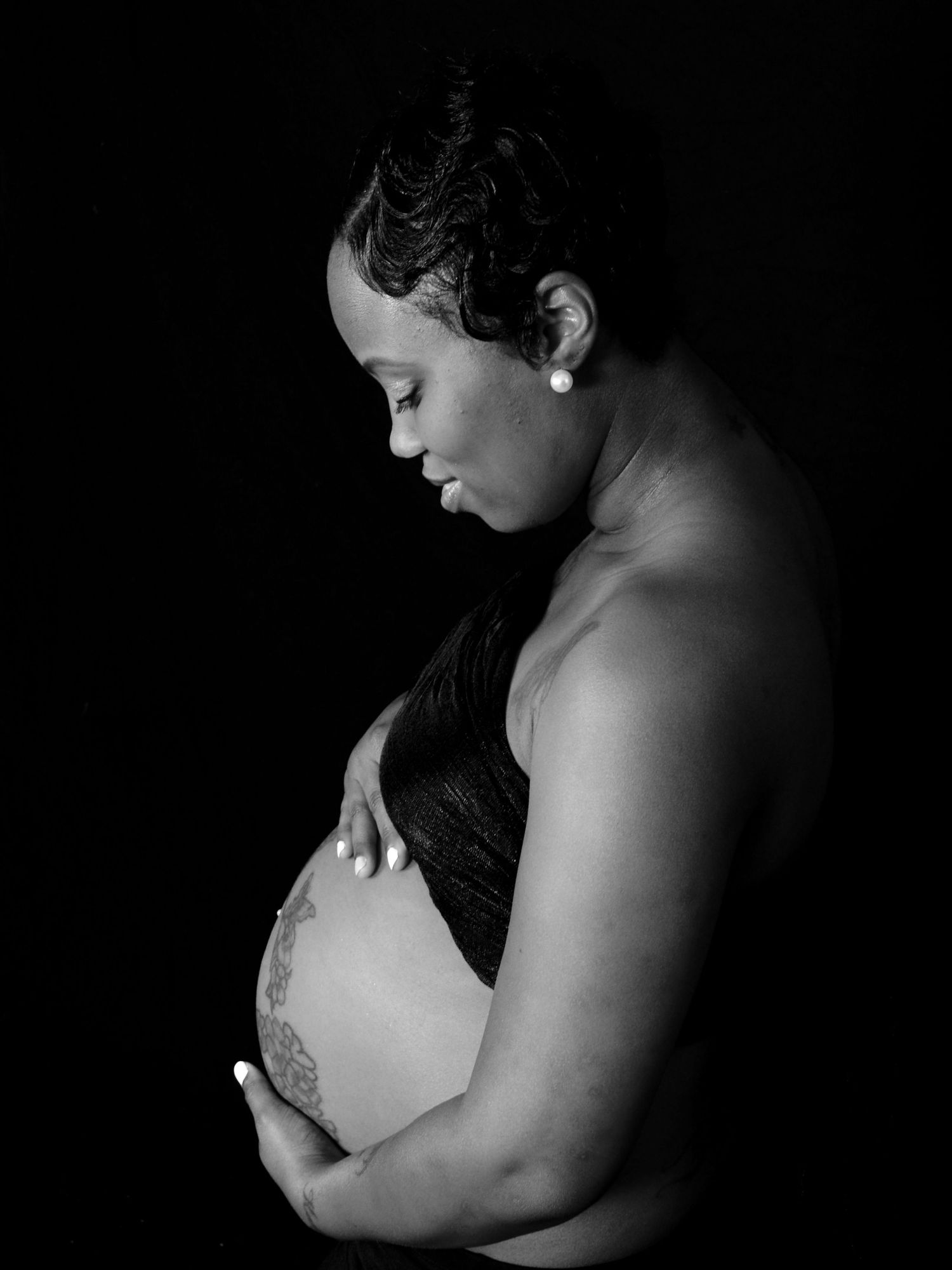Elegant black and white profile portrait of pregnant woman cradling her baby bump in darkness.