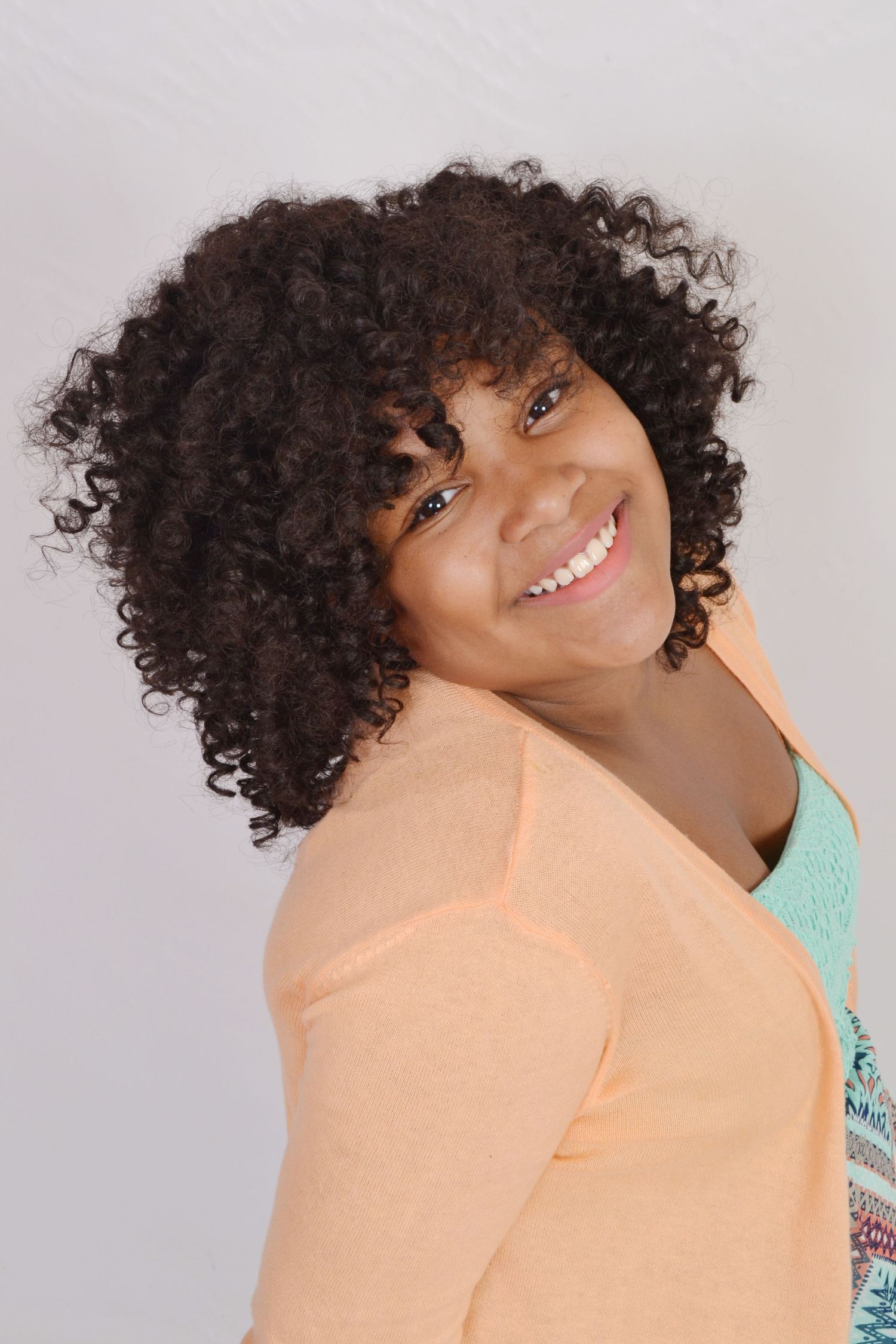 Smiling young girl with curly hair wearing peach cardigan poses against light gray background.