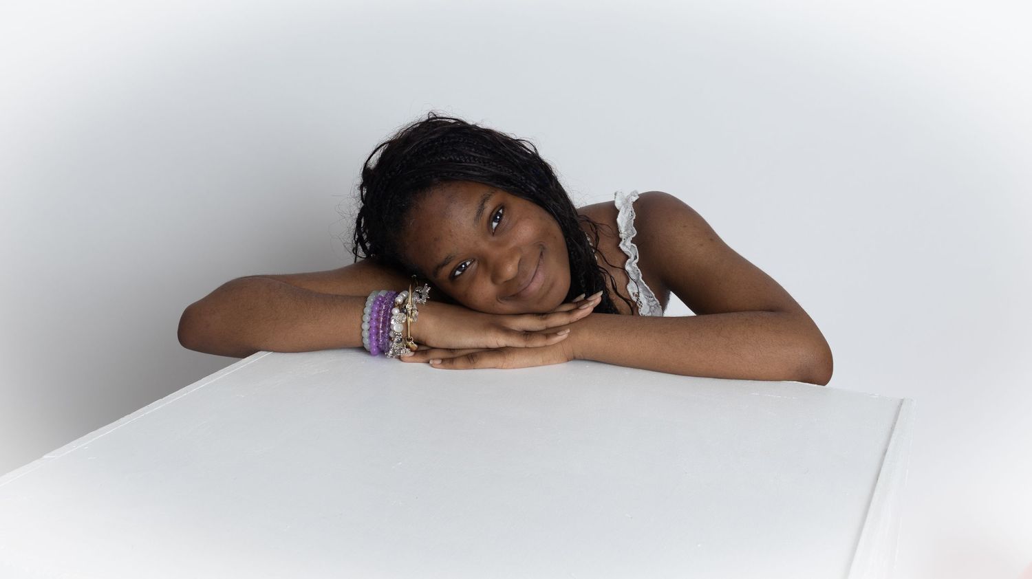 Young Black girl with braids and colorful bracelets rests her chin on hands at a white table against a light background.