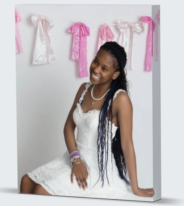 Young girl in white lace dress with braids smiles, seated before pink ribbon decorations on white background.