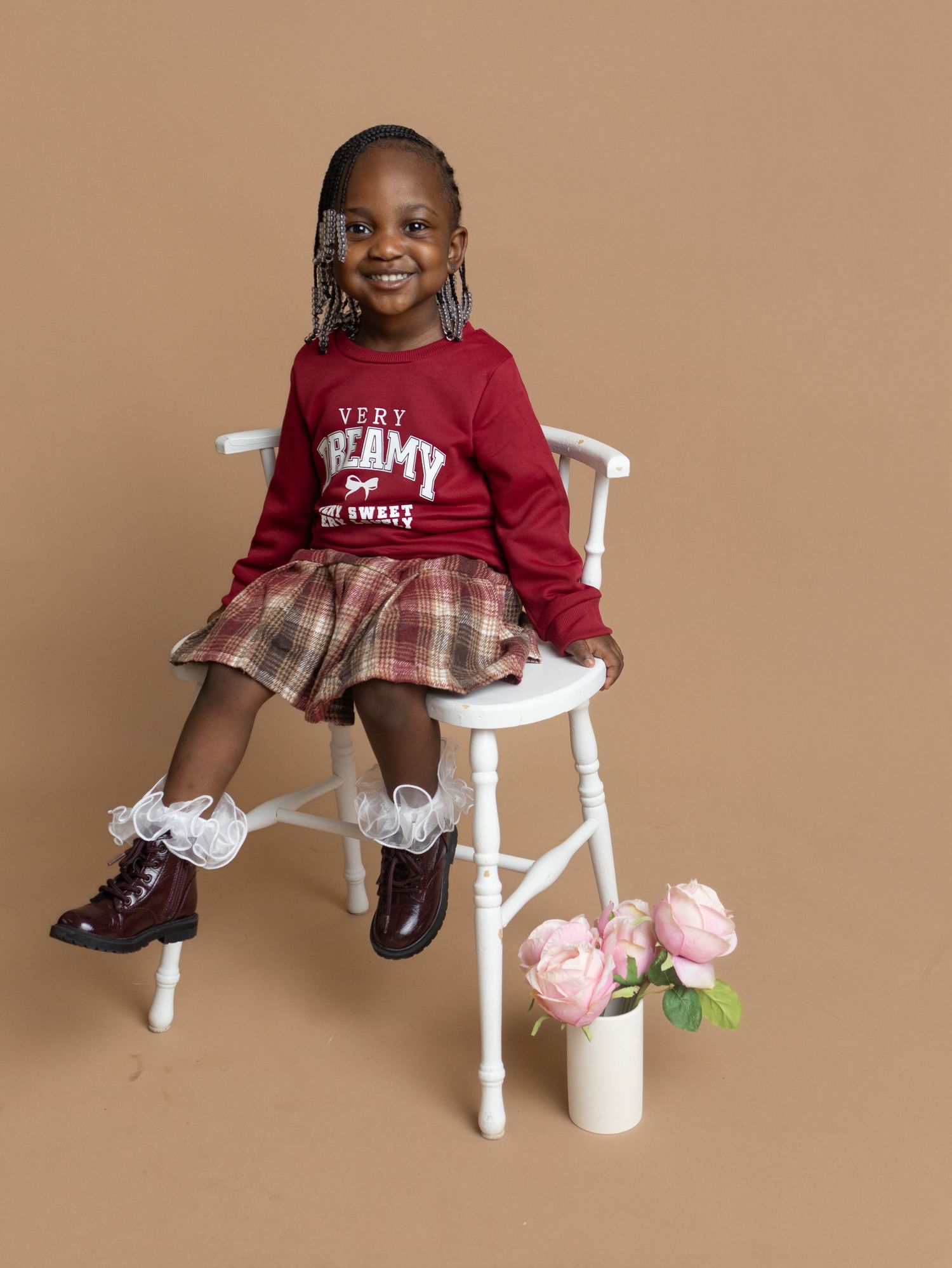Young girl in red sweater and plaid skirt sits on white chair, smiling, with pink roses nearby.