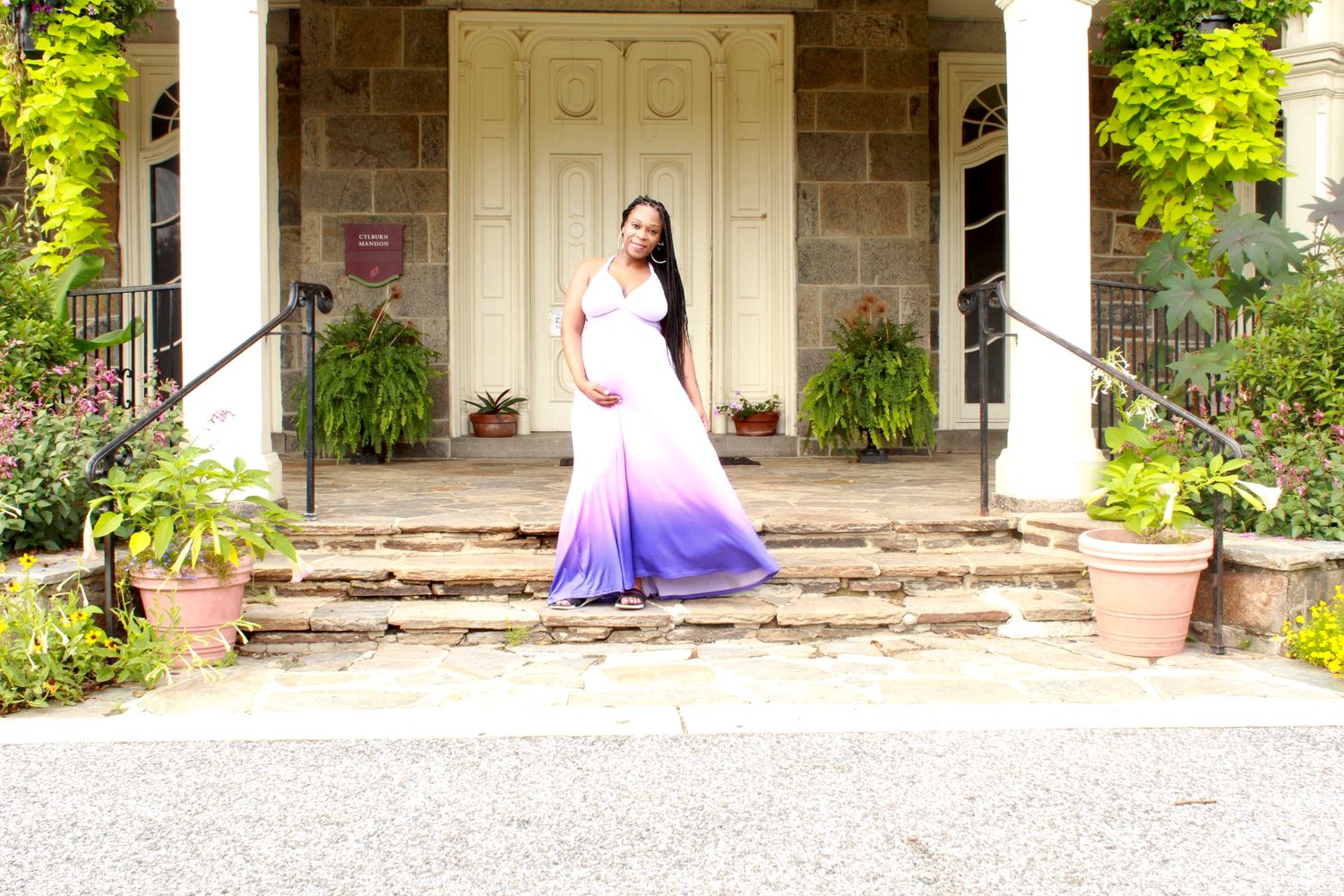 A pregnant woman in a white-to-purple ombre gown stands elegantly on steps outside a historic stone building.