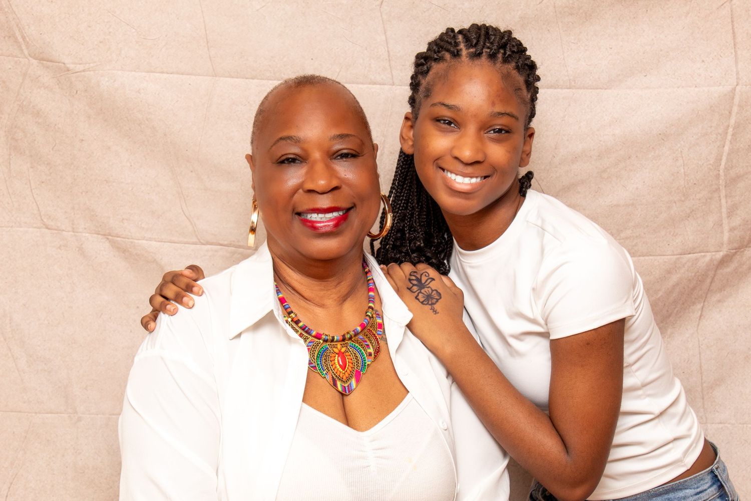 Two smiling women pose together, one older with a colorful necklace and one younger with braids, both wearing white tops.