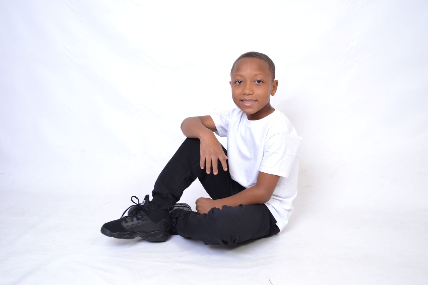 Young boy in white tee and black pants sits casually on white floor smiling at camera.
