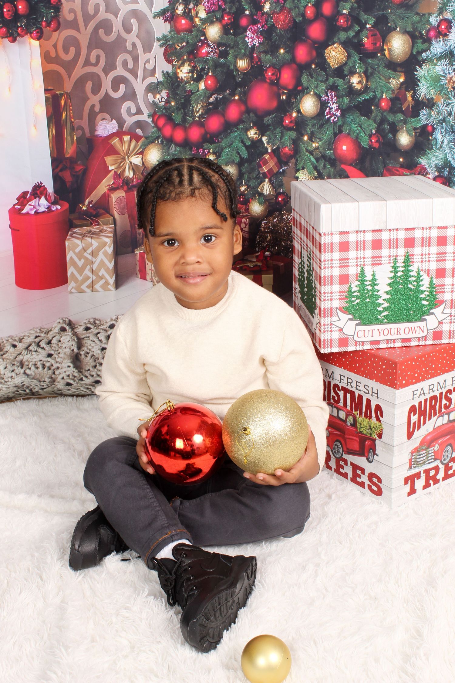 A young boy in a cream sweater sits cross-legged holding red and gold ornaments in front of a Christmas tree.