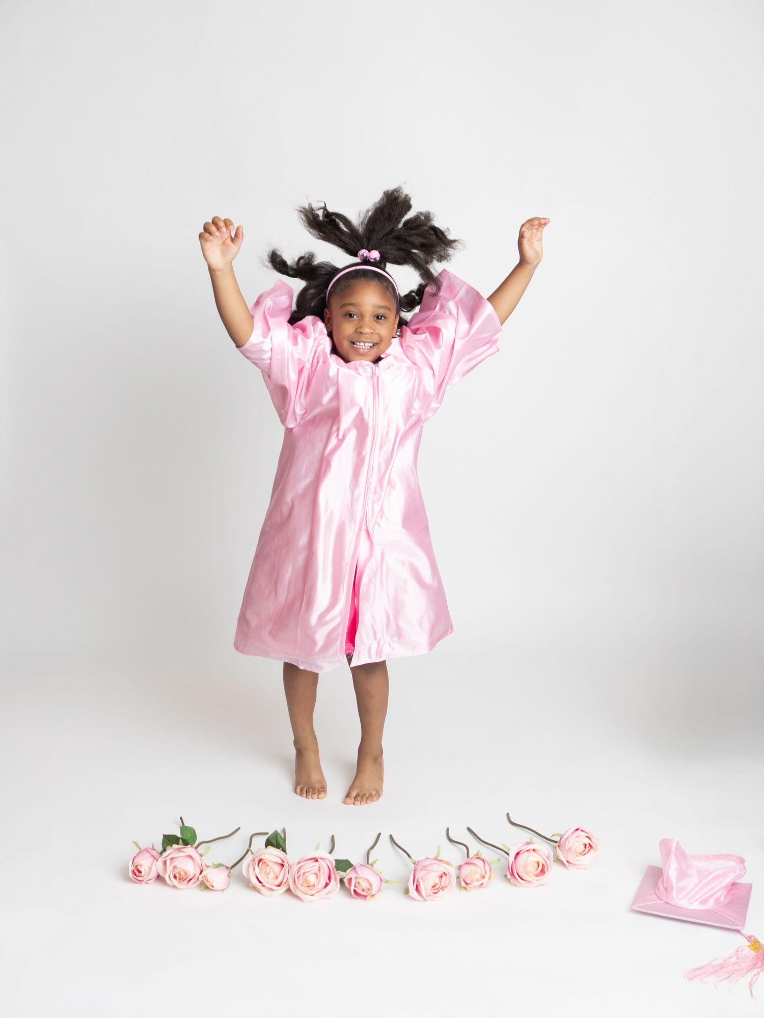 Joyful young girl jumping in pink graduation gown with roses on floor below her.