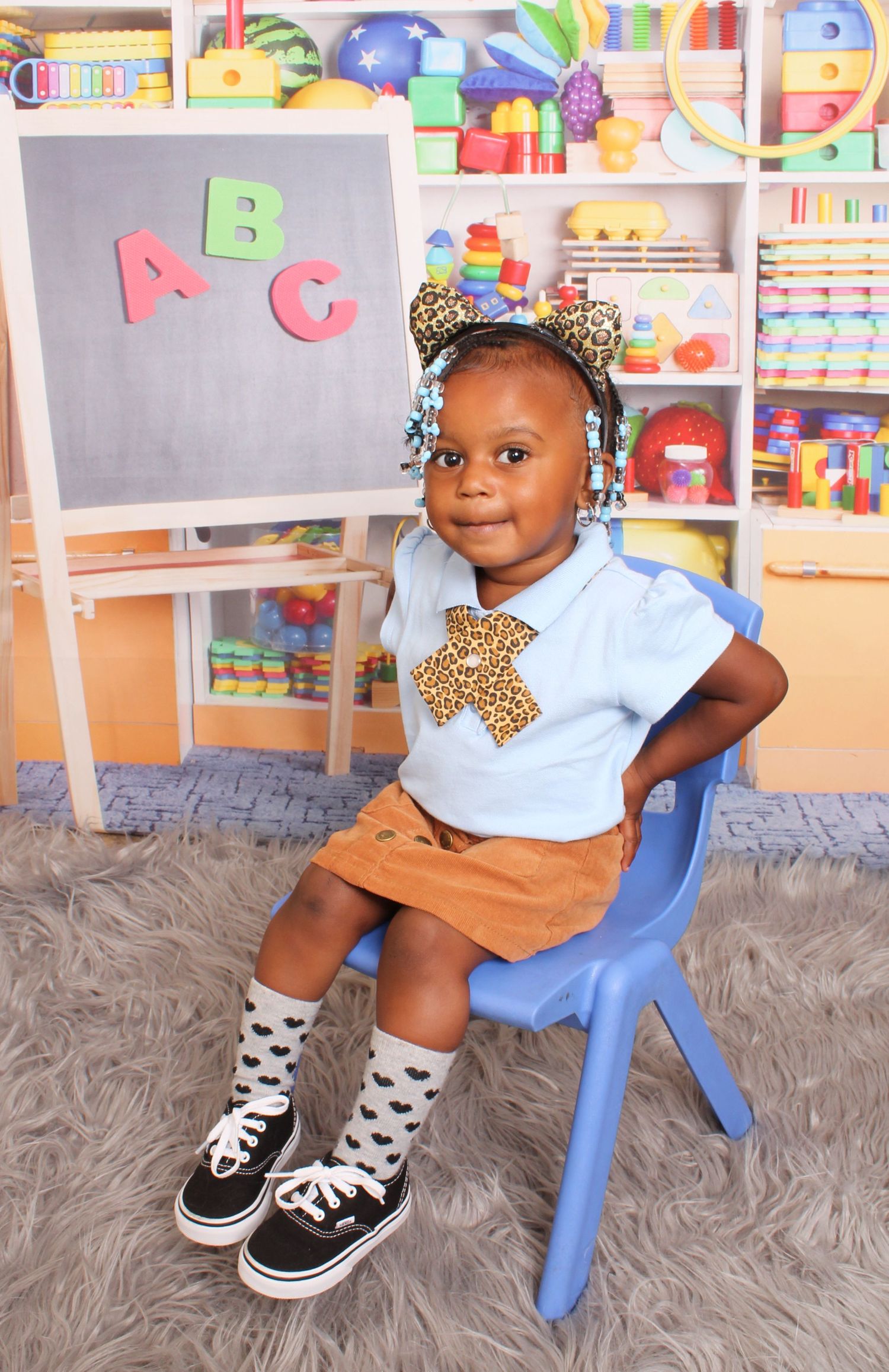 A stylish toddler in a blue polo and leopard print bow sits in a blue chair before an ABC chalkboard.