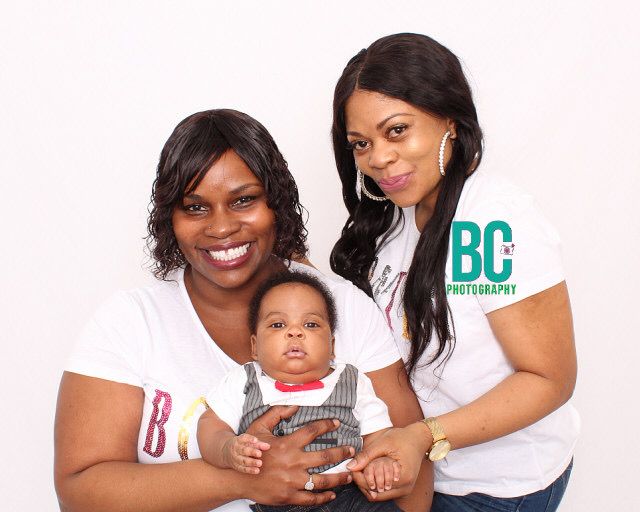Two women smile proudly while holding a baby boy in matching BC Photography white shirts.