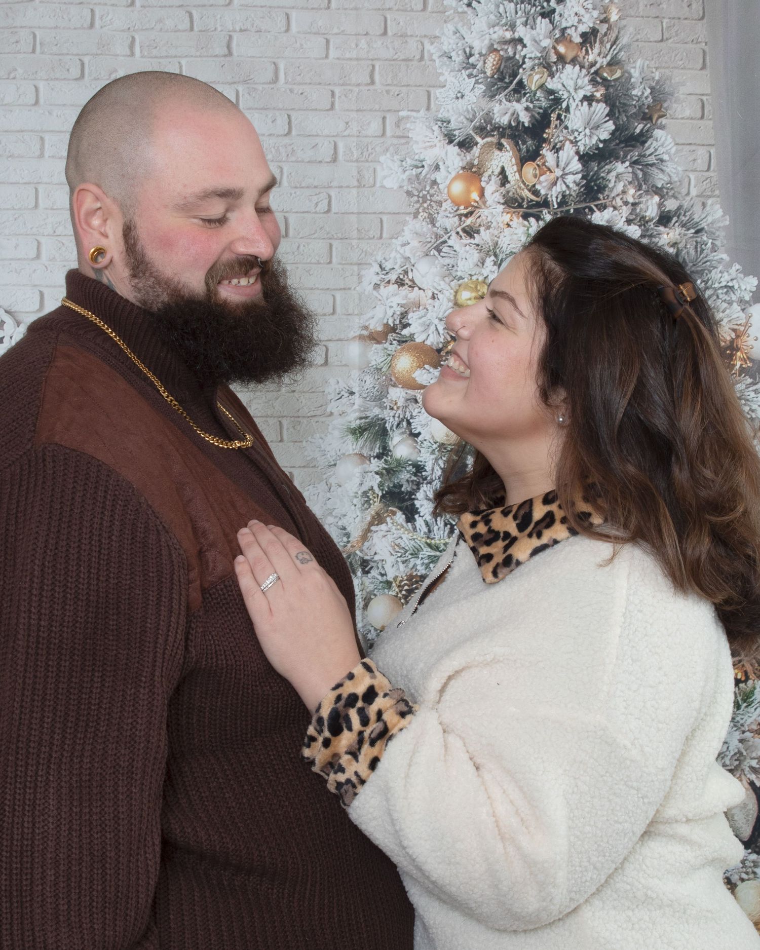 A bearded man and smiling woman share a tender moment in front of a flocked Christmas tree.