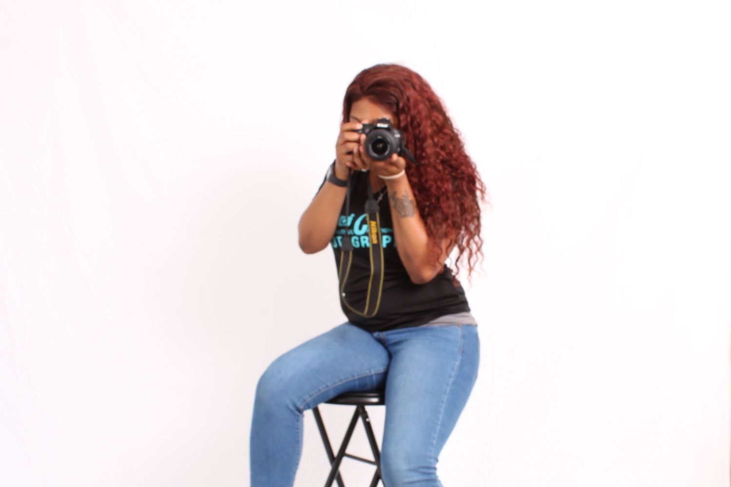 Woman with red hair sitting on stool holding DSLR camera up to face against white background.