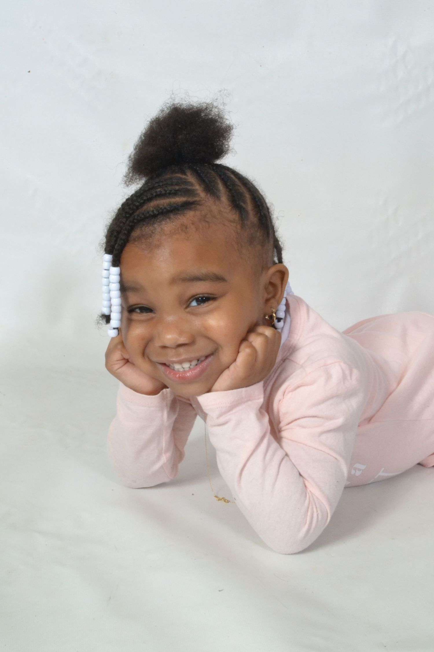 Cute little girl in pink outfit lies on white surface smiling with hands on cheeks.