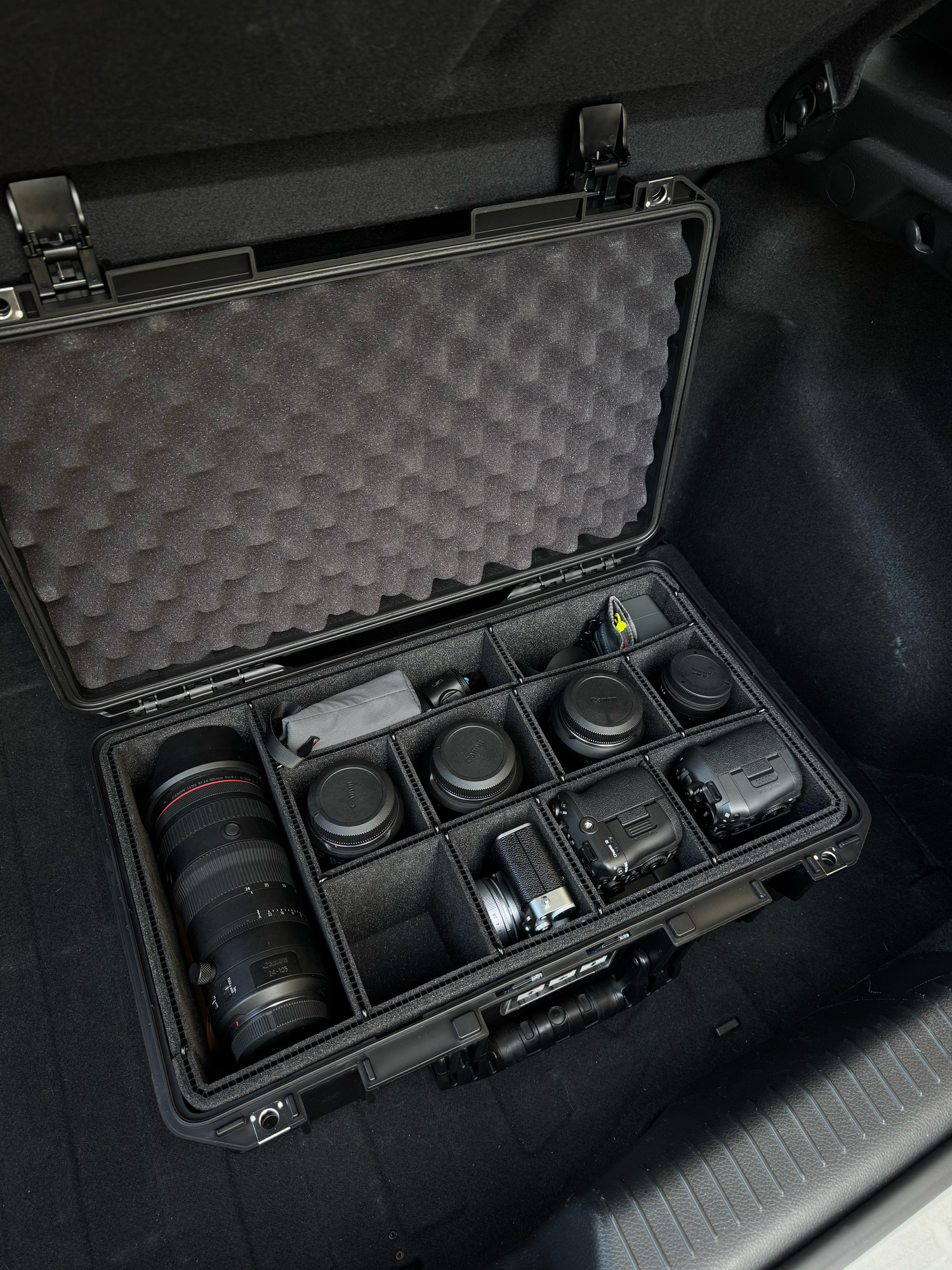 Professional camera equipment organized in a black Pelican hard case with foam dividers, stored in a vehicle trunk.