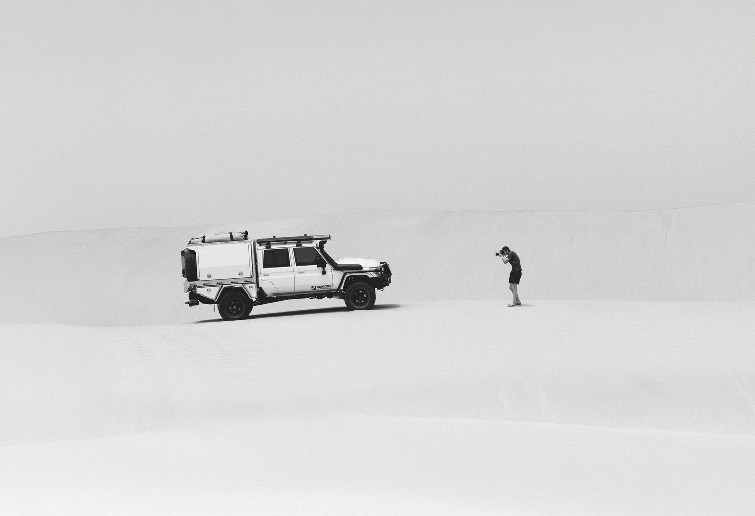 Black and white photo of a 4WD vehicle and photographer on vast white sand dunes in a remote desert landscape.