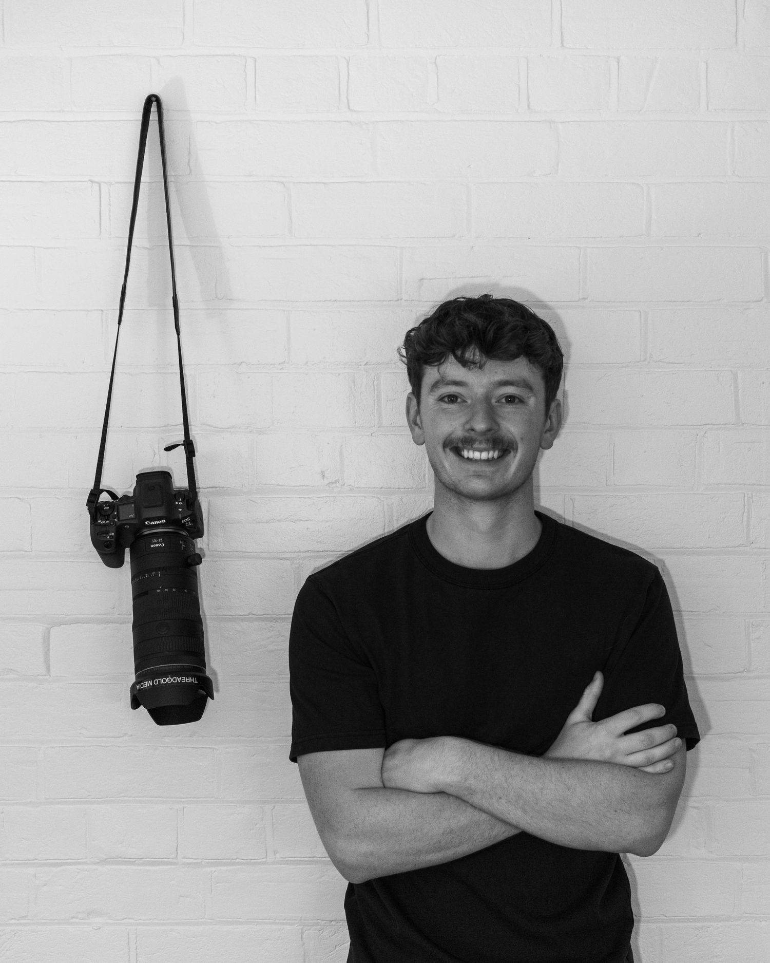 Young man with mustache smiling, arms crossed, standing beside a DSLR camera on white brick wall.