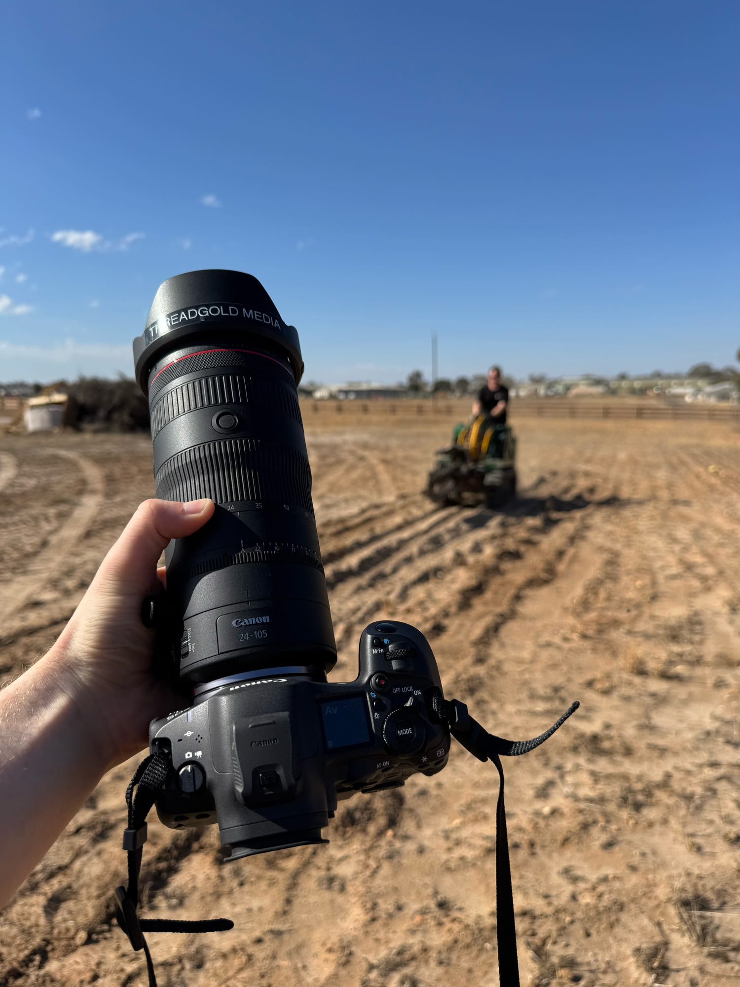 A hand holds a Canon DSLR camera with telephoto lens in a dirt field, with a farmer on a tractor in the background.