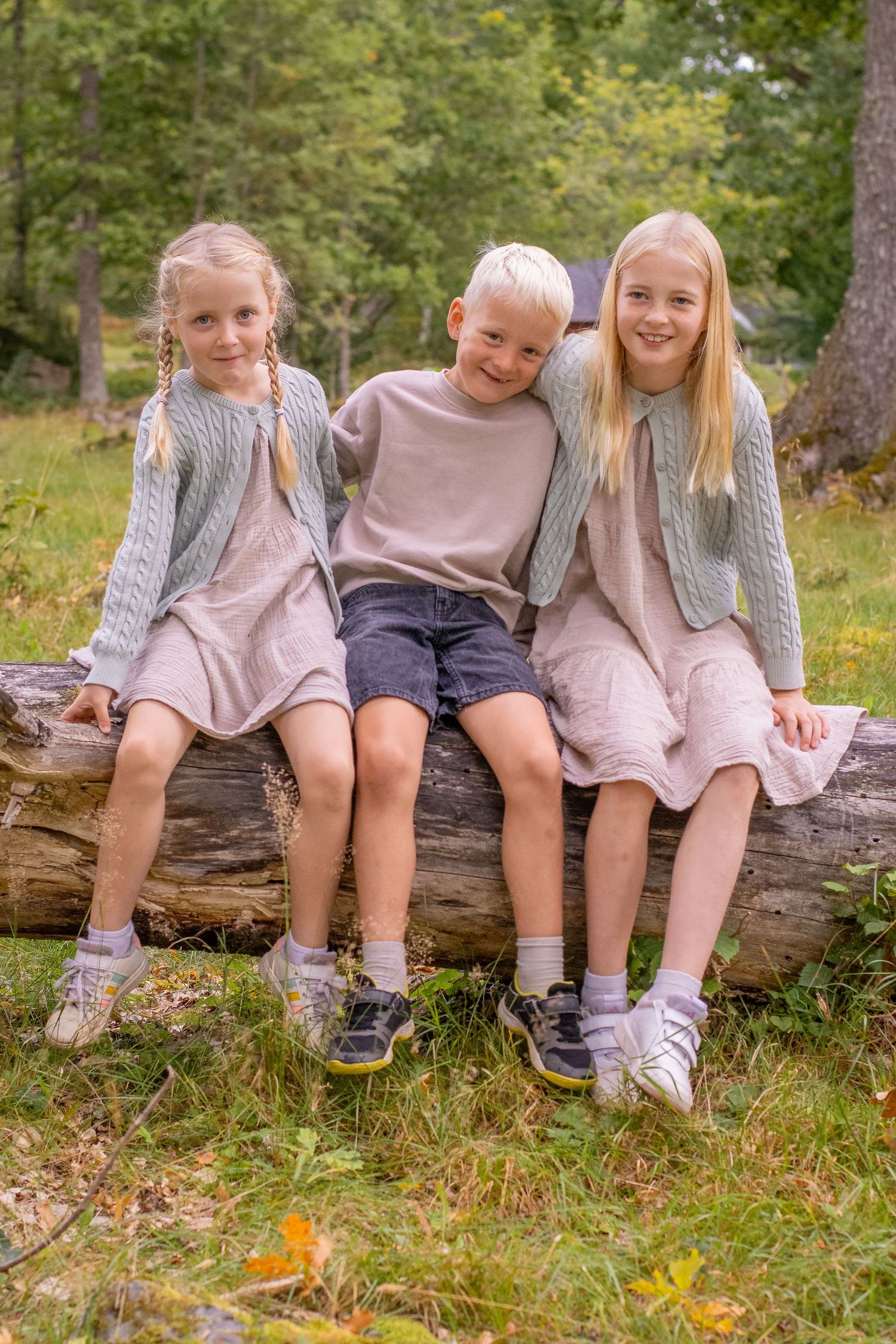 Tre barn sitter på en liggande stock i skogsmiljö, en pojke i mitten och en flicka på varje sida. Barnen tittar in i kameran och ler.