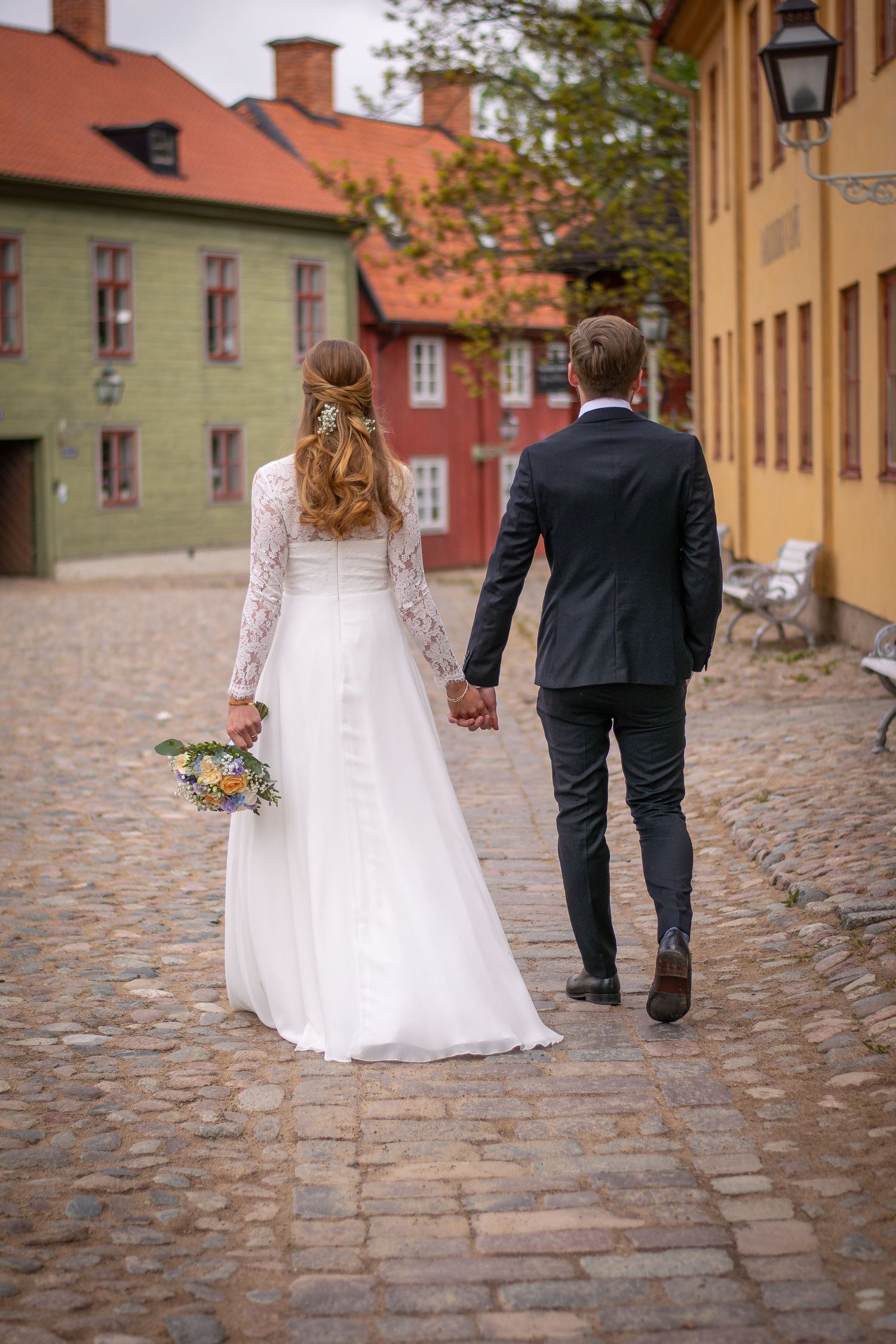 Brudpar gåendes hand i hand med ryggen mot kameran, på en gammal stengata med färstarka hus bakgrunden.