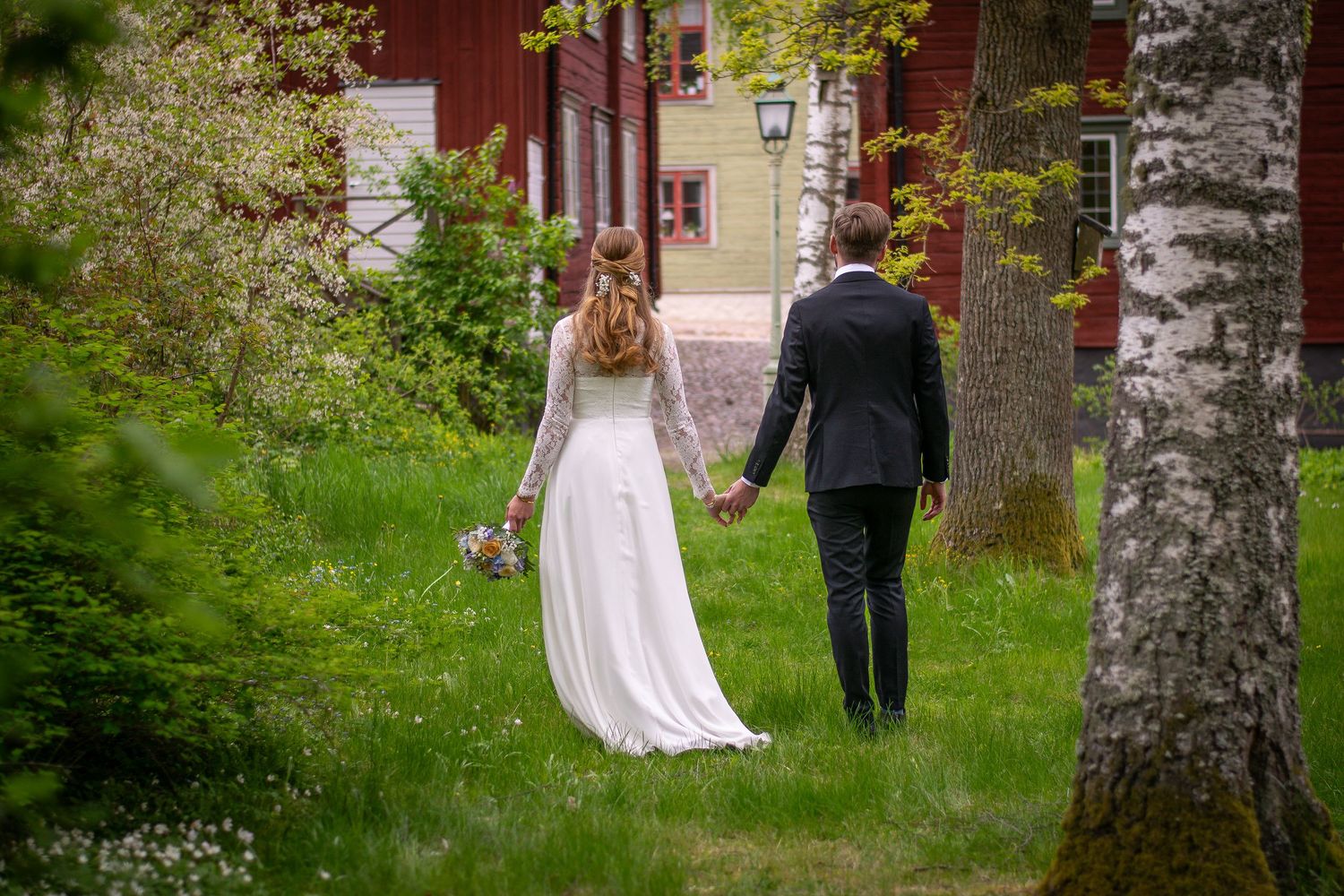 Brudpar gåendes hand i hand med ryggen mot kameran, i en grönskande omgivning och med röda hus i bakgrunden.