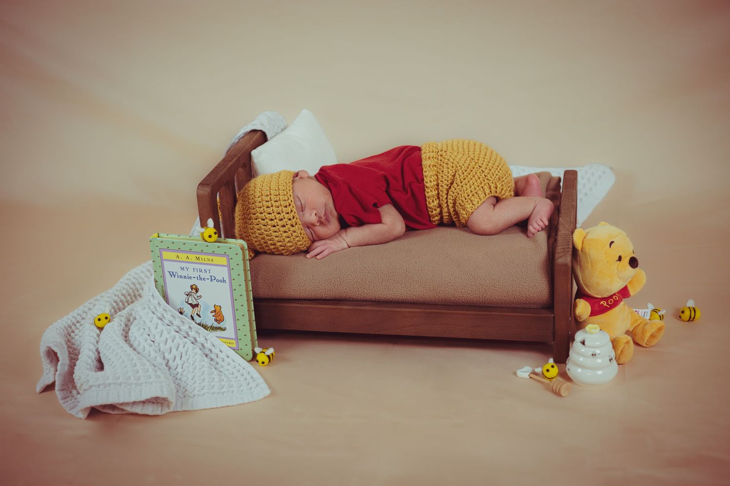 Newborn baby dressed as Winnie the Pooh sleeping on tiny wooden bed with stuffed animal and books.