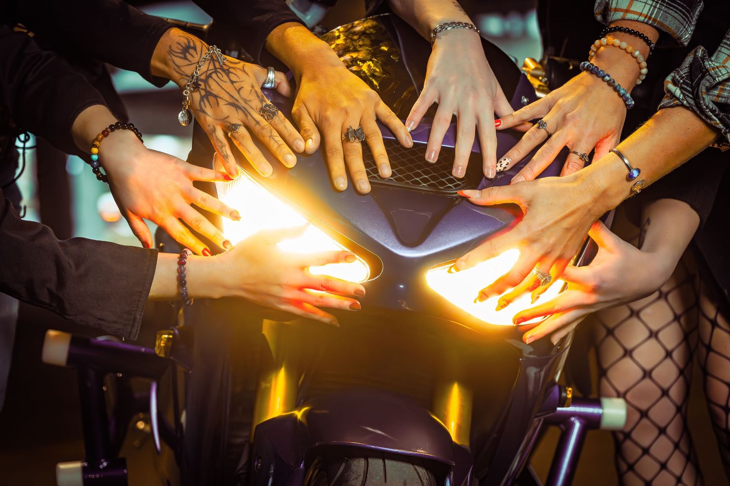 Multiple hands reach toward a motorcycle headlight glowing warmly in a dark setting.