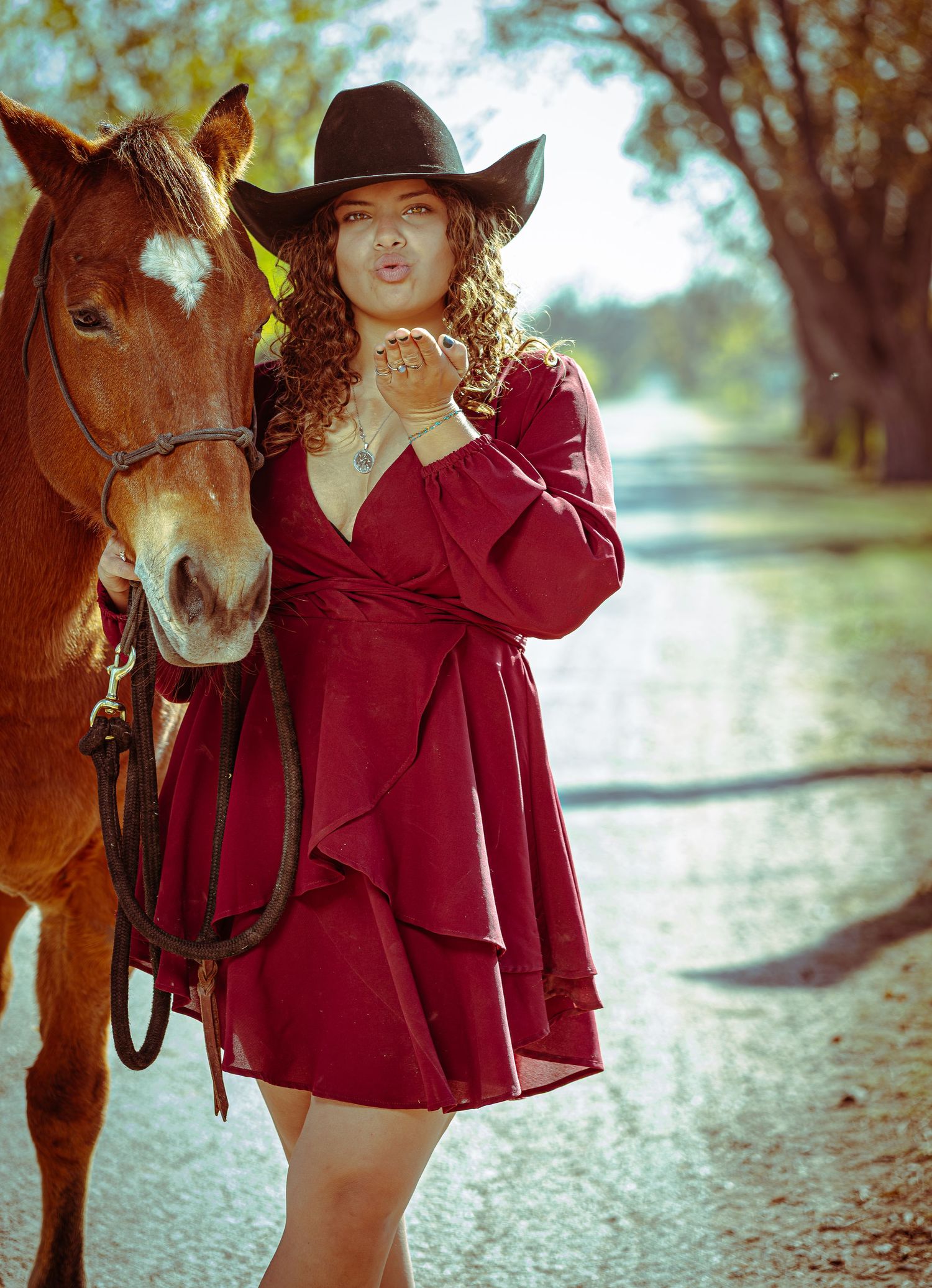 A person in a red jacket and cowboy hat poses with a brown horse on a sunlit country path.