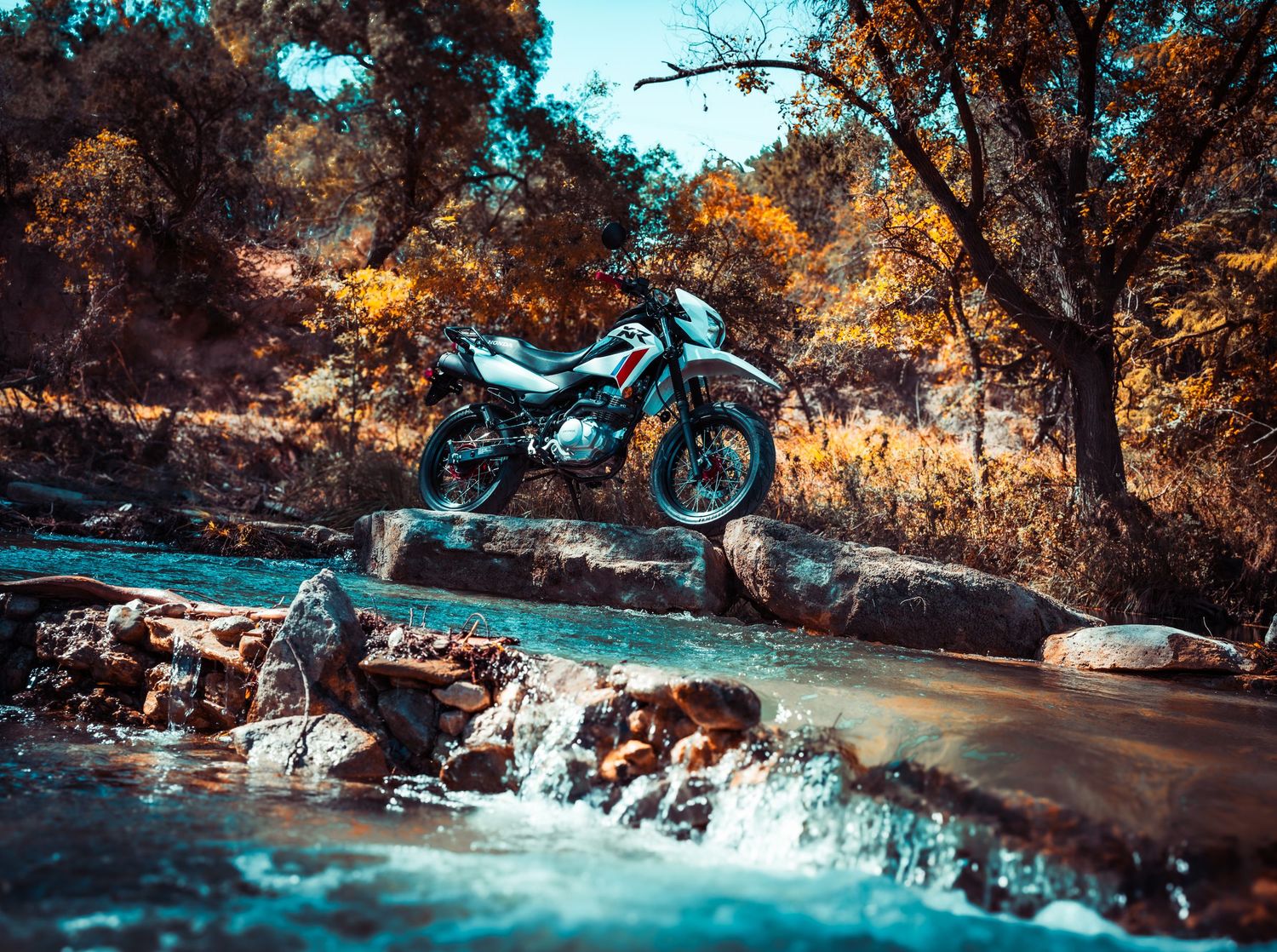 Dual sport motorcycle positioned on rocks beside flowing stream with orange autumn trees in background.