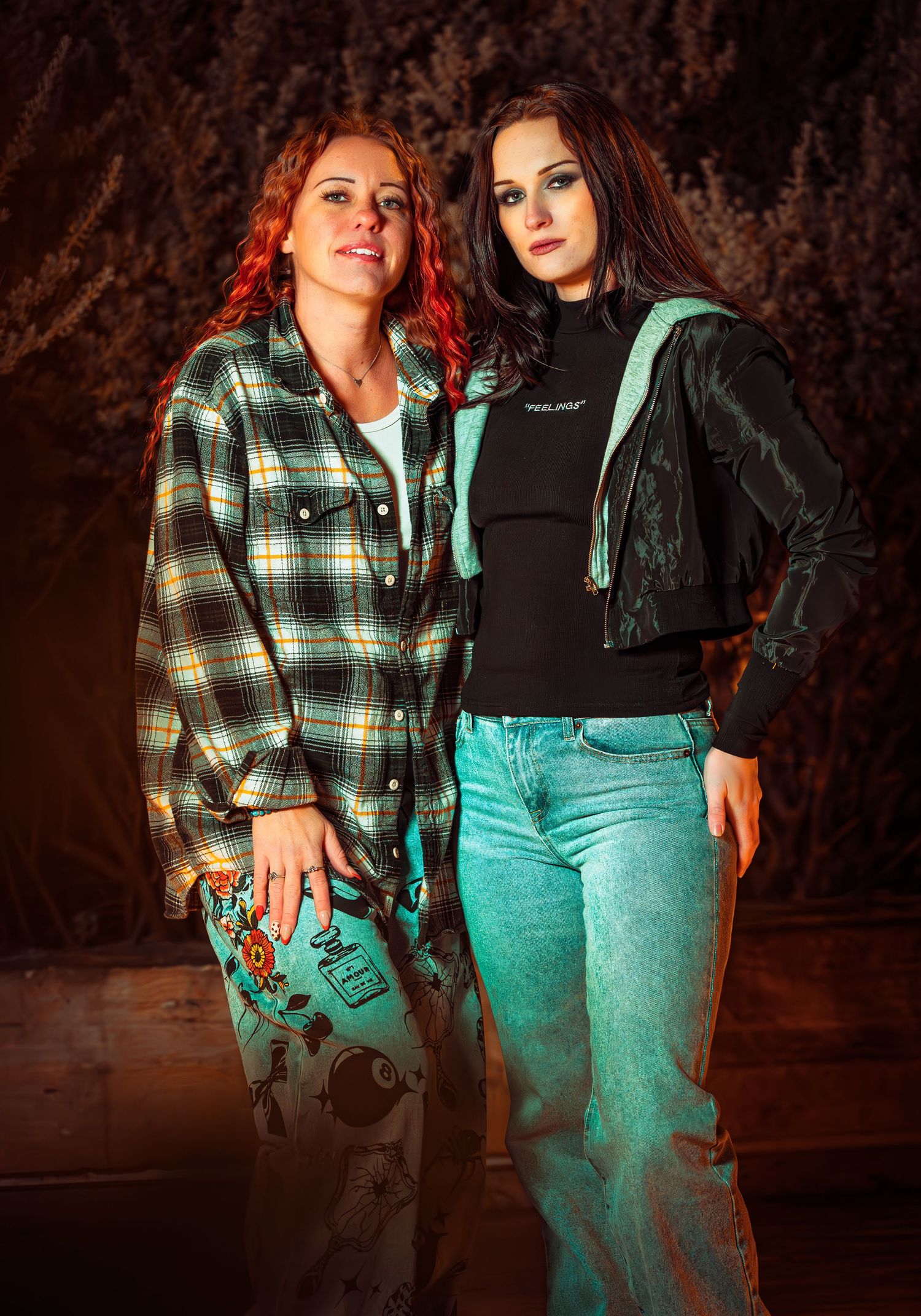 Two people pose together wearing casual outfits with plaid flannel and denim against a dark textured background.