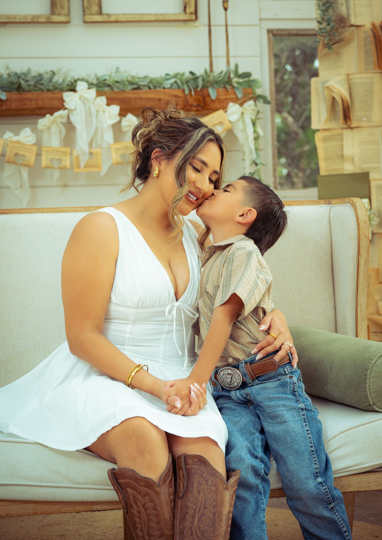 A woman in a white dress and cowboy boots sits on a couch as a young boy gives her a sweet kiss on the cheek.