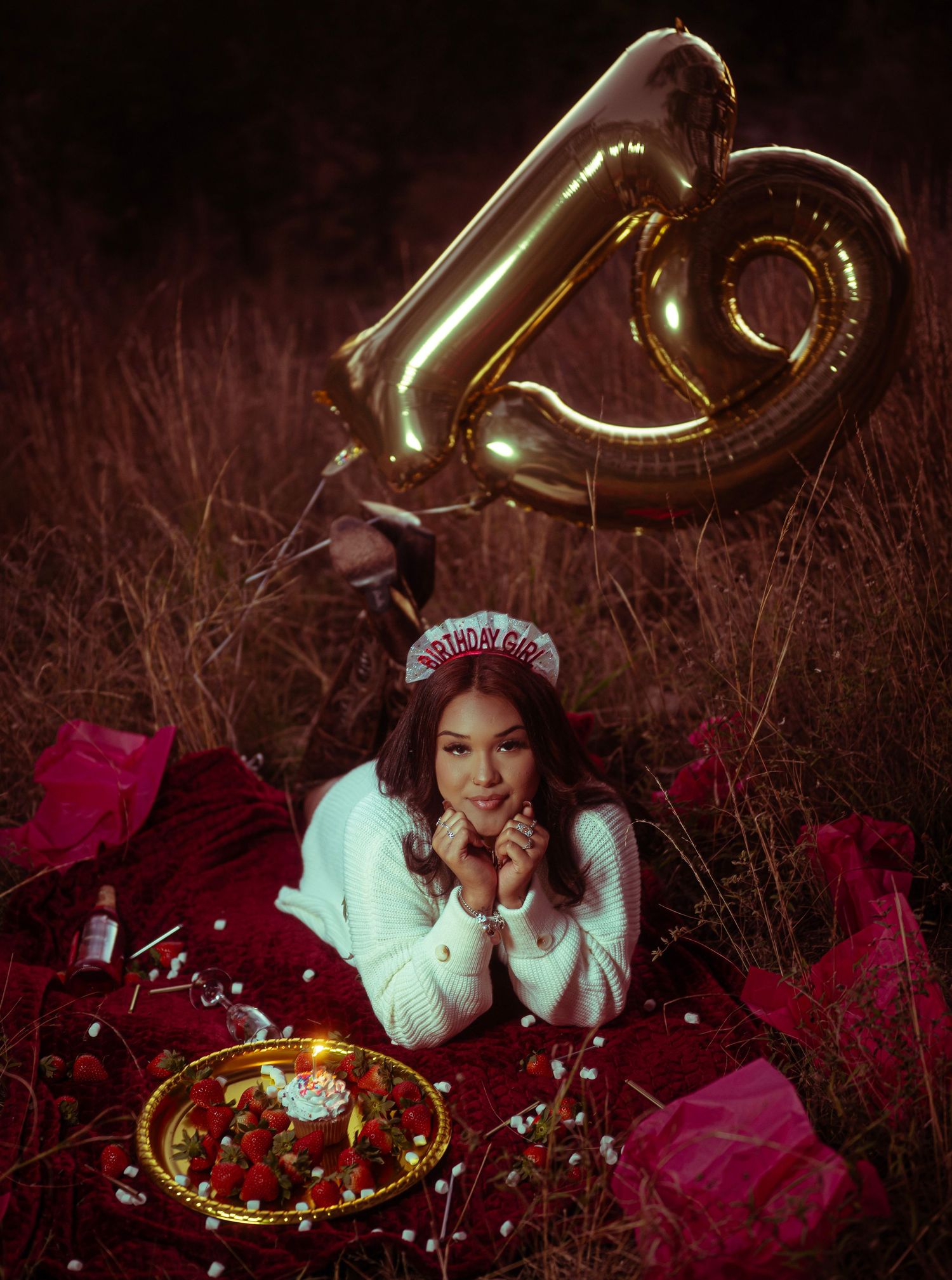 A dreamy winter scene with gold balloons and red decor featuring someone lying in grass wearing a white sweater and crown.