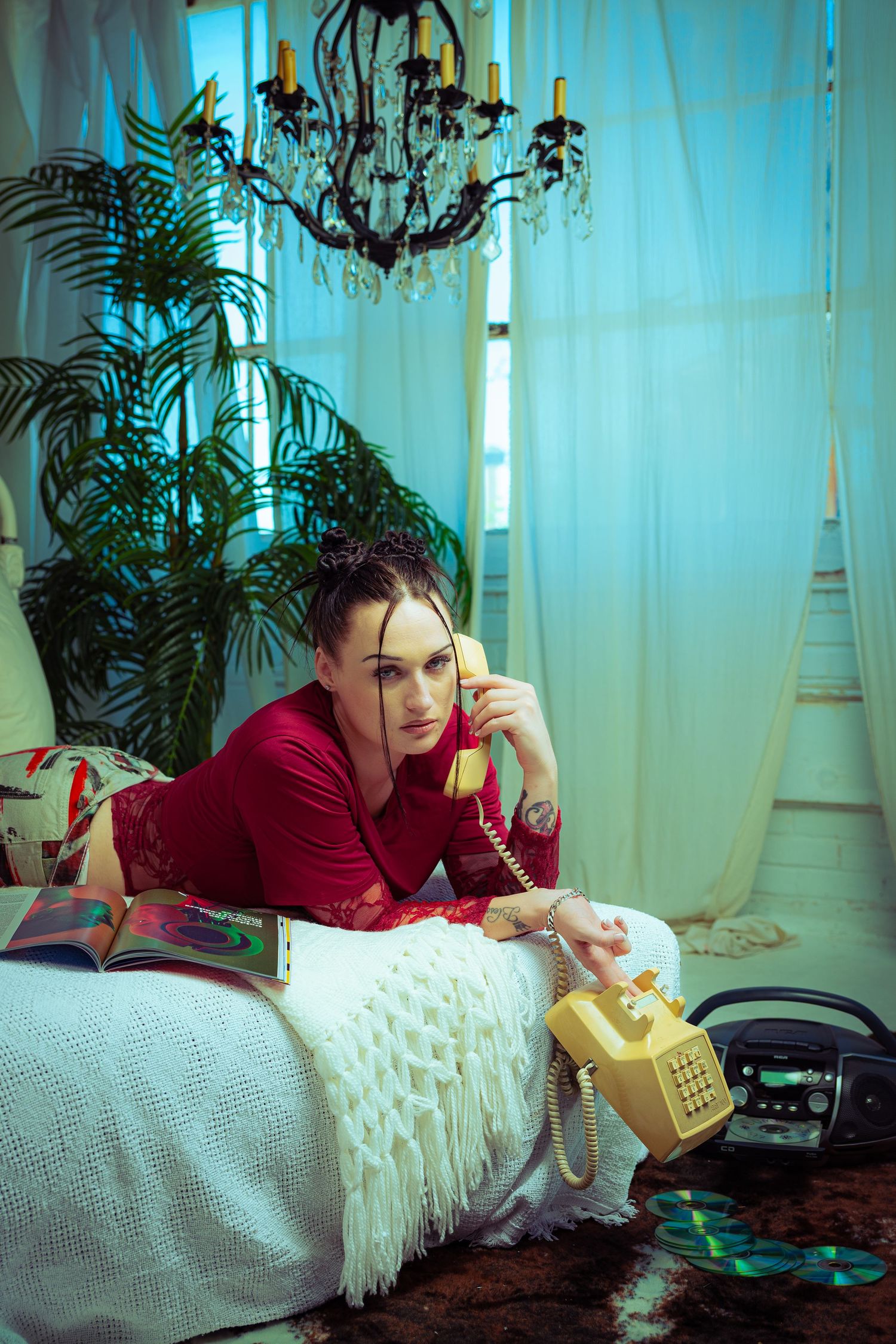 Woman in red top lying on bed, talking on vintage yellow rotary phone in retro-styled room with chandelier.