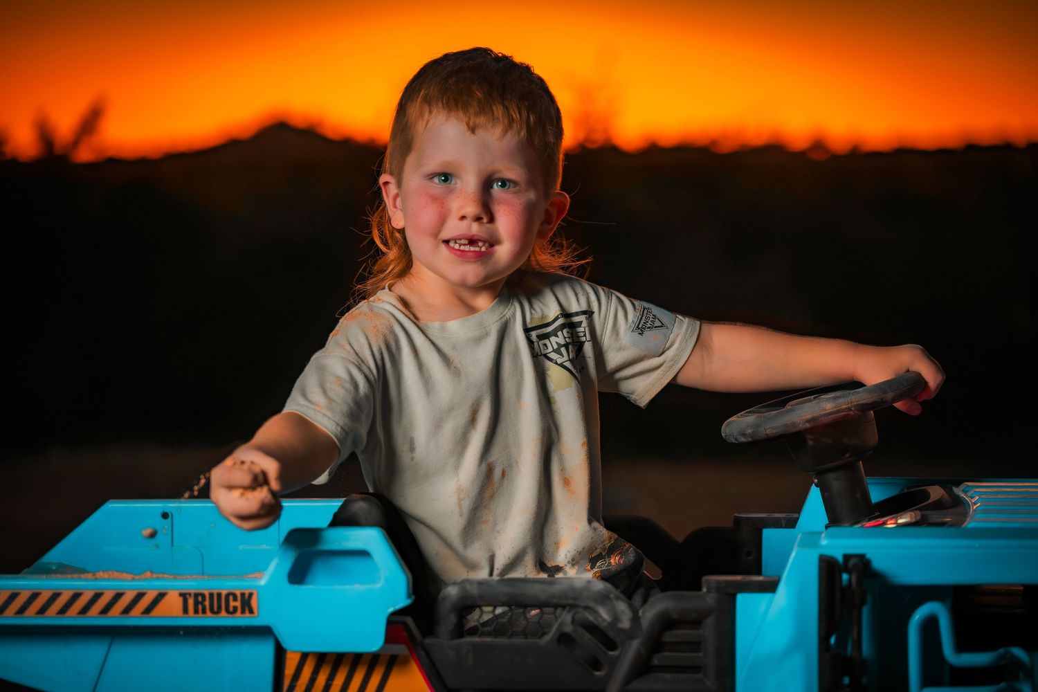 Child playing with blue toy truck against vibrant sunset background.