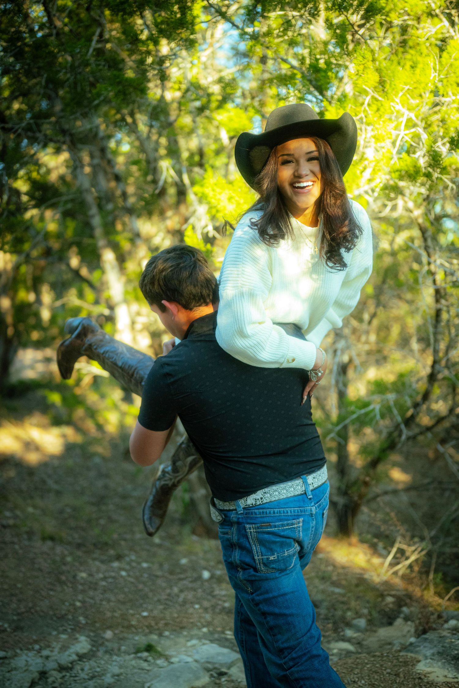 A person wearing a white sweater, jeans and cowboy hat poses playfully in a sunlit wooded outdoor setting.