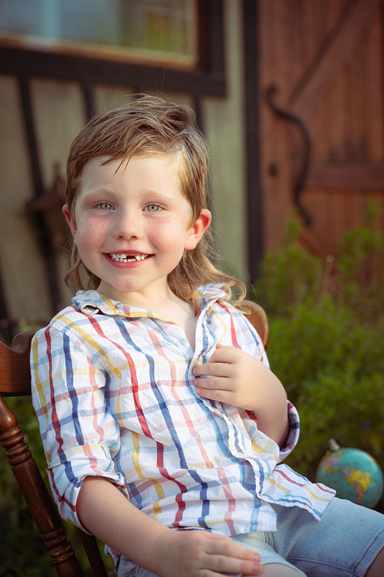Smiling young child with missing front tooth sits in wooden chair wearing colorful striped shirt outdoors in warm sunlight.